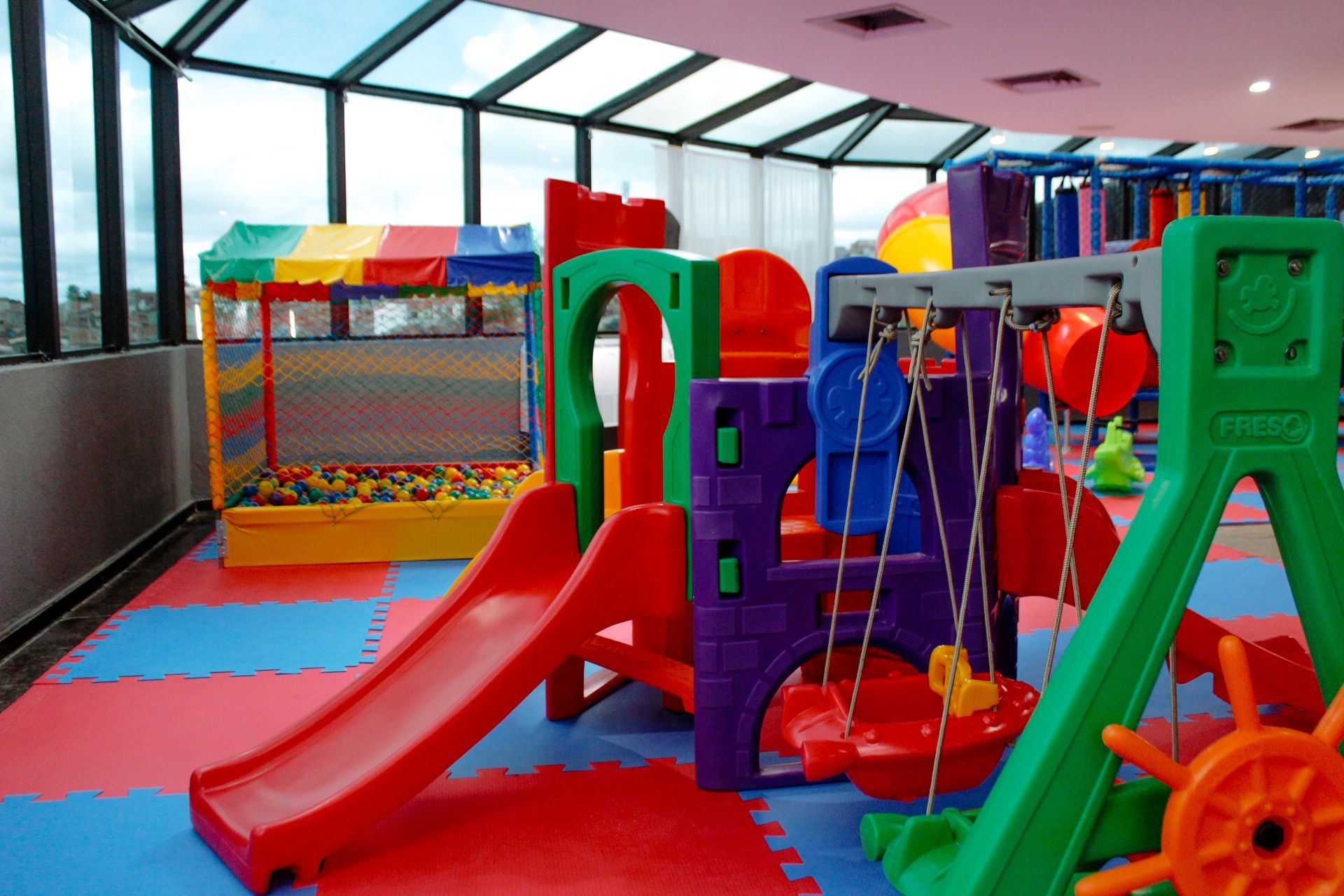 Parque infantil coberto com estrutura de brinquedos colorida, piscina de bolinhas e balanços em piso acolchoado.