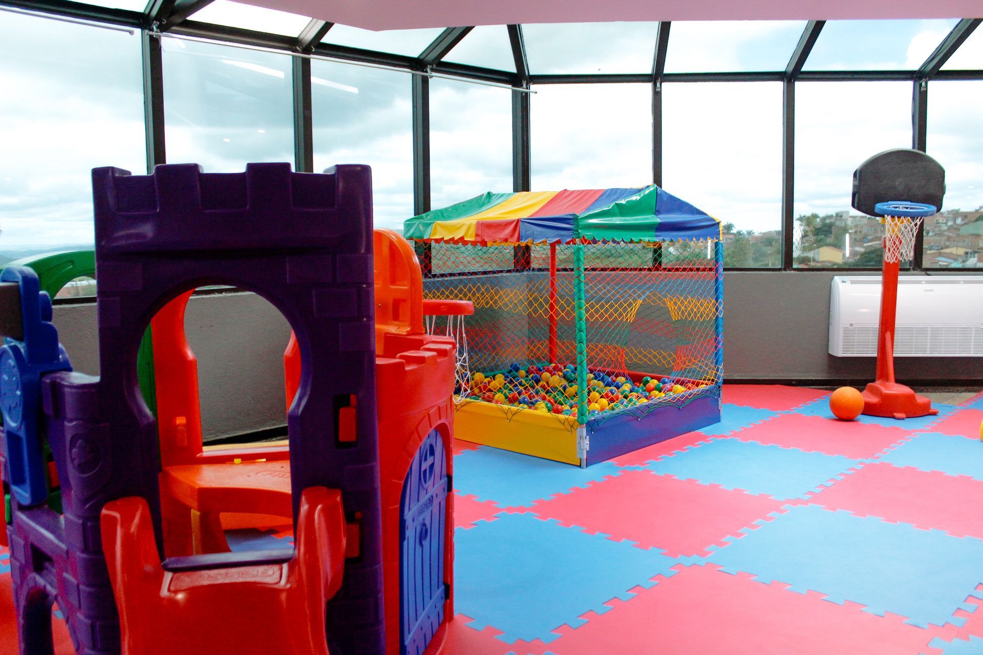 Área de recreação infantil interna com castelo colorido, piscina de bolinhas, cesta de basquete e vista para a janela.