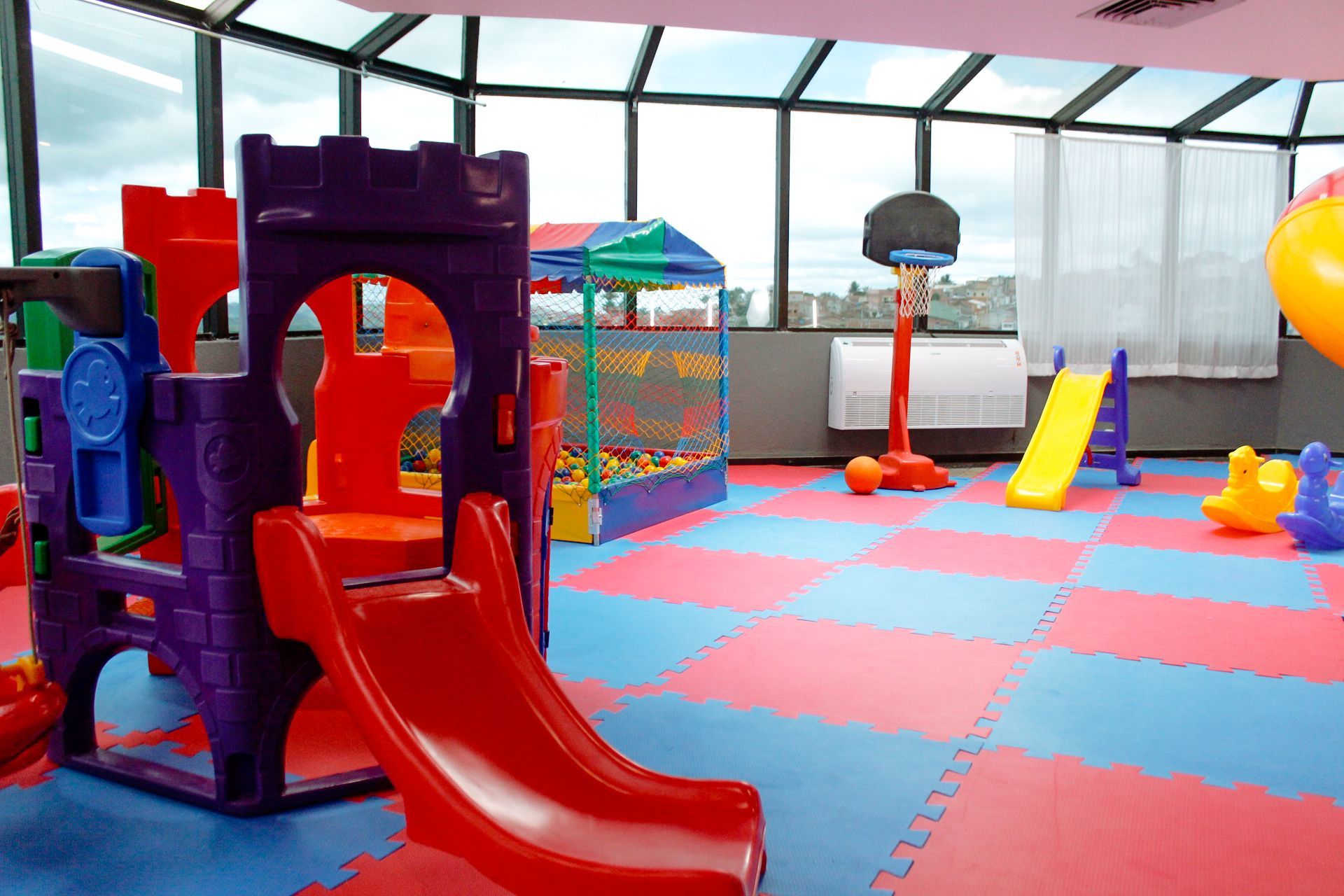 Sala de jogos com estrutura de escalada em formato de castelo, escorregador, piscina de bolinhas, cesta de basquete e tapetes coloridos no chão.