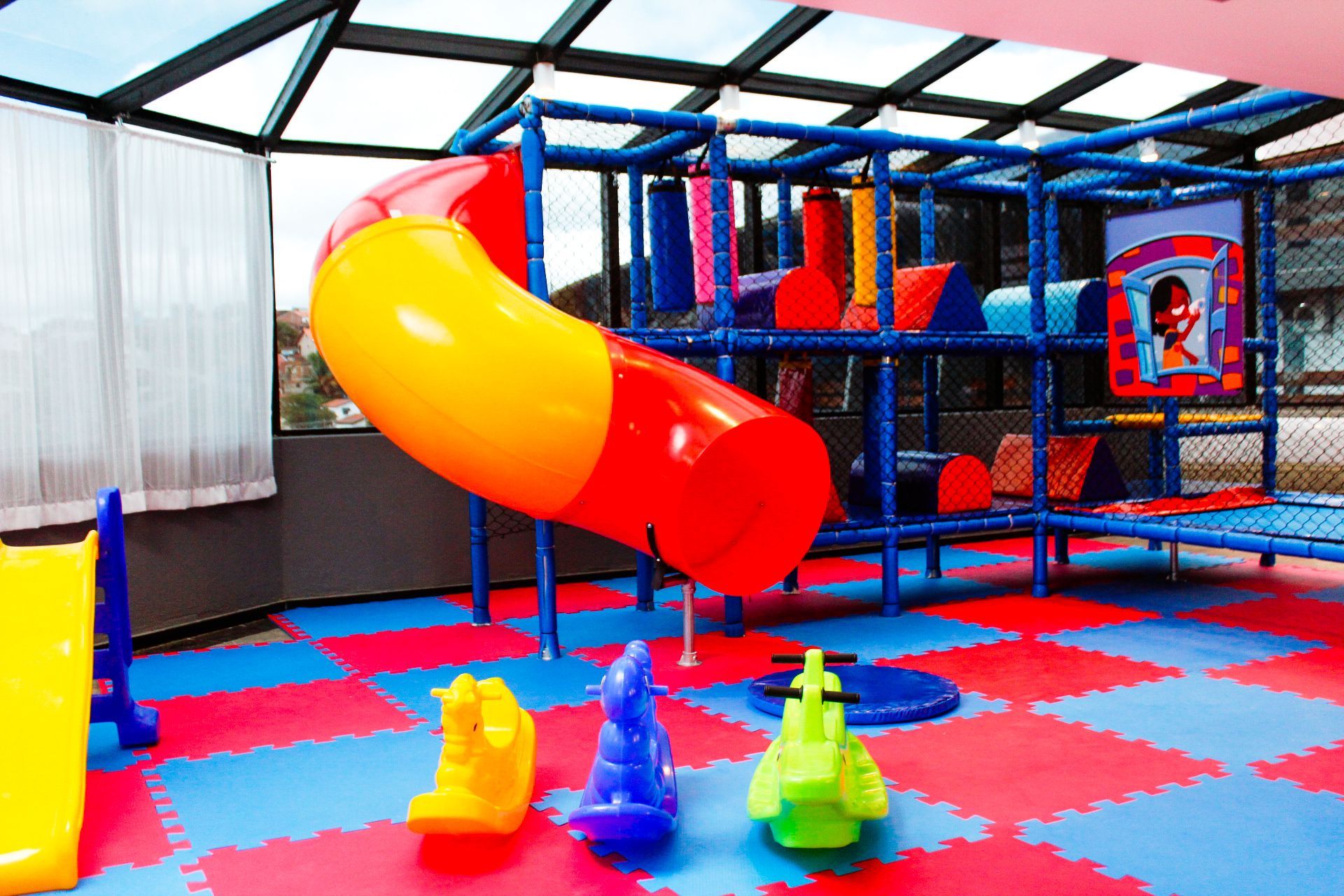Área de recreação com escorregador azul, vermelho e amarelo, tapetes e equipamentos de playground.