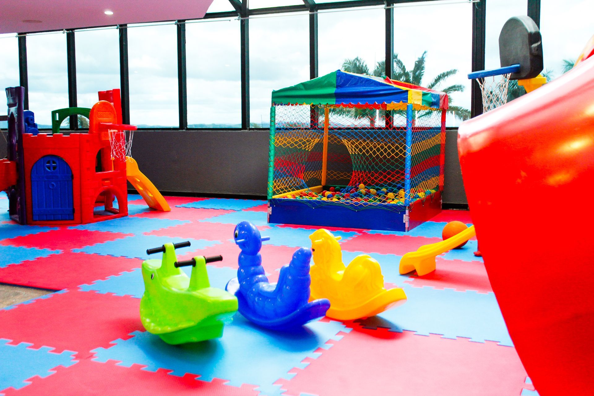 Área de recreação infantil interna com estruturas de brinquedo vermelhas, azuis, verdes e amarelas, piscina de bolinhas e cesta de basquete, com grandes janelas.