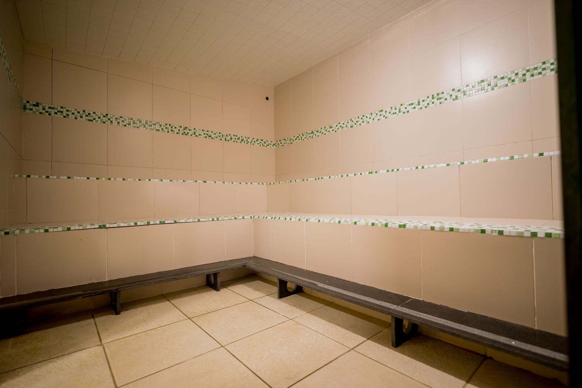 Sala vazia com piso de azulejo, prateleiras e bancos. Paredes bege-claras, detalhes decorativos verdes, bancos cinza.