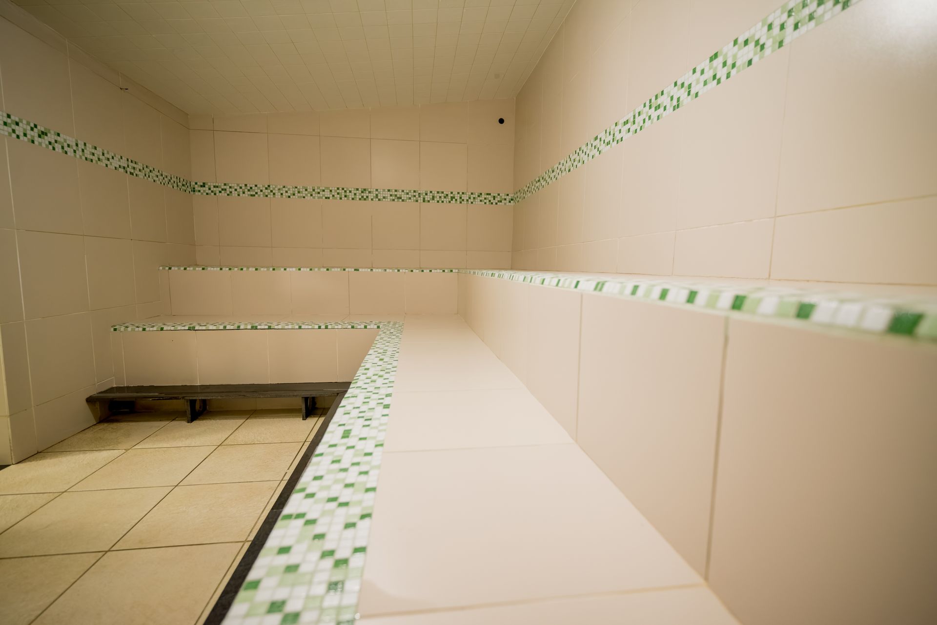 Sala de vapor vazia com paredes, bancos e piso revestidos de azulejos, com detalhes em verde.