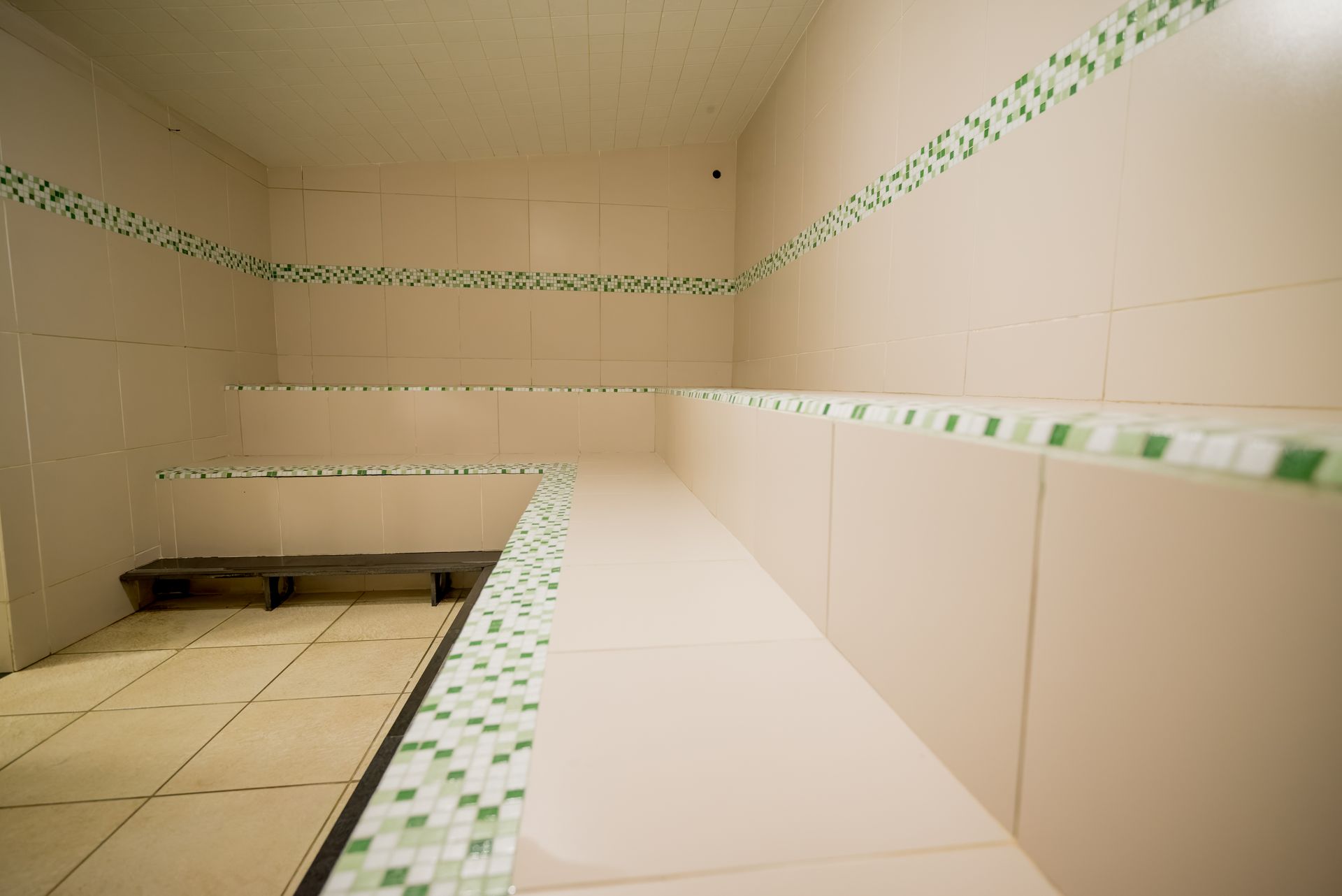 Interior da sala de vapor revestida com azulejos, bancos embutidos, detalhes em mosaico verde e um banco de madeira.