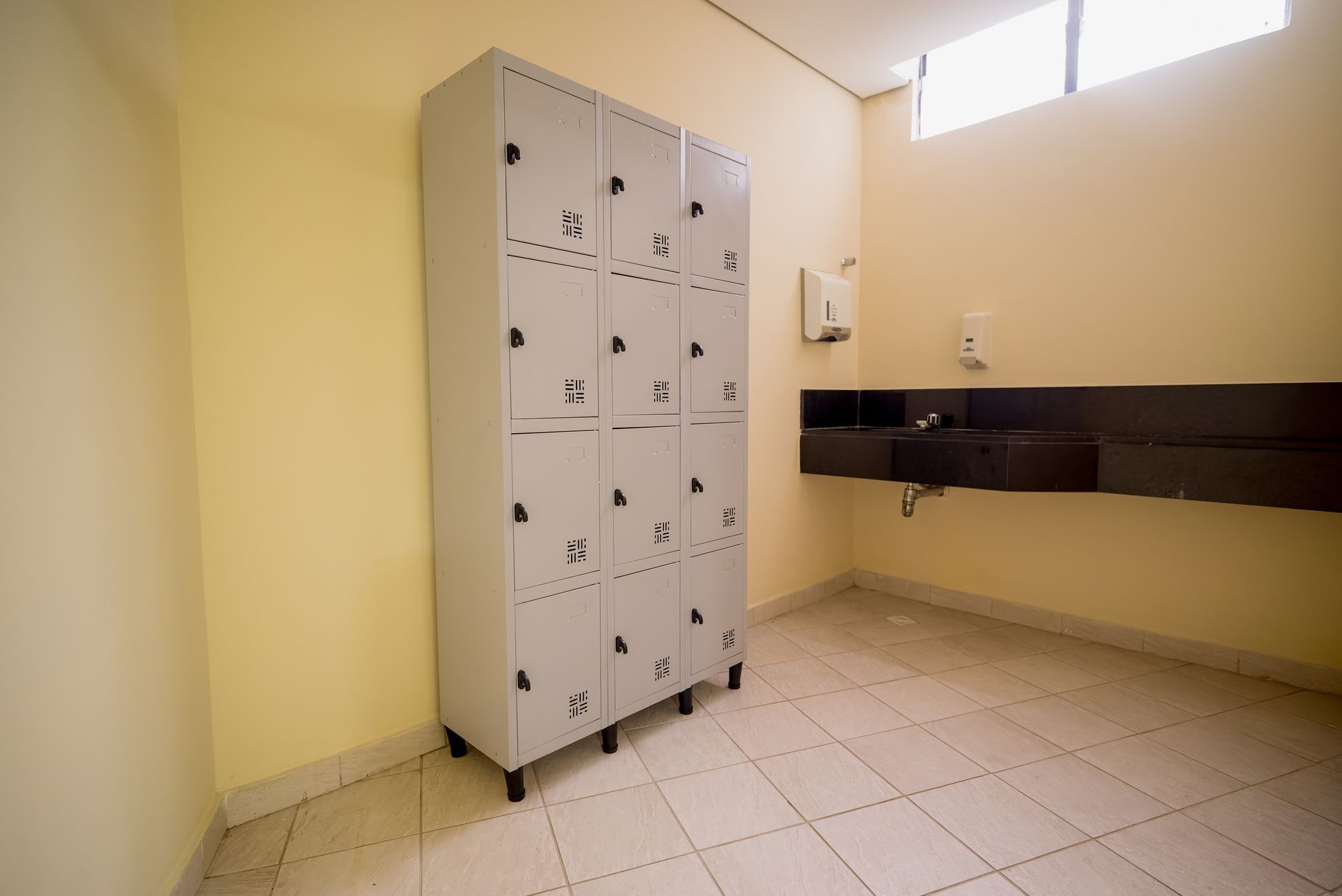 Um vestiário com armários cinzentos, uma bancada escura com pia e uma janela.