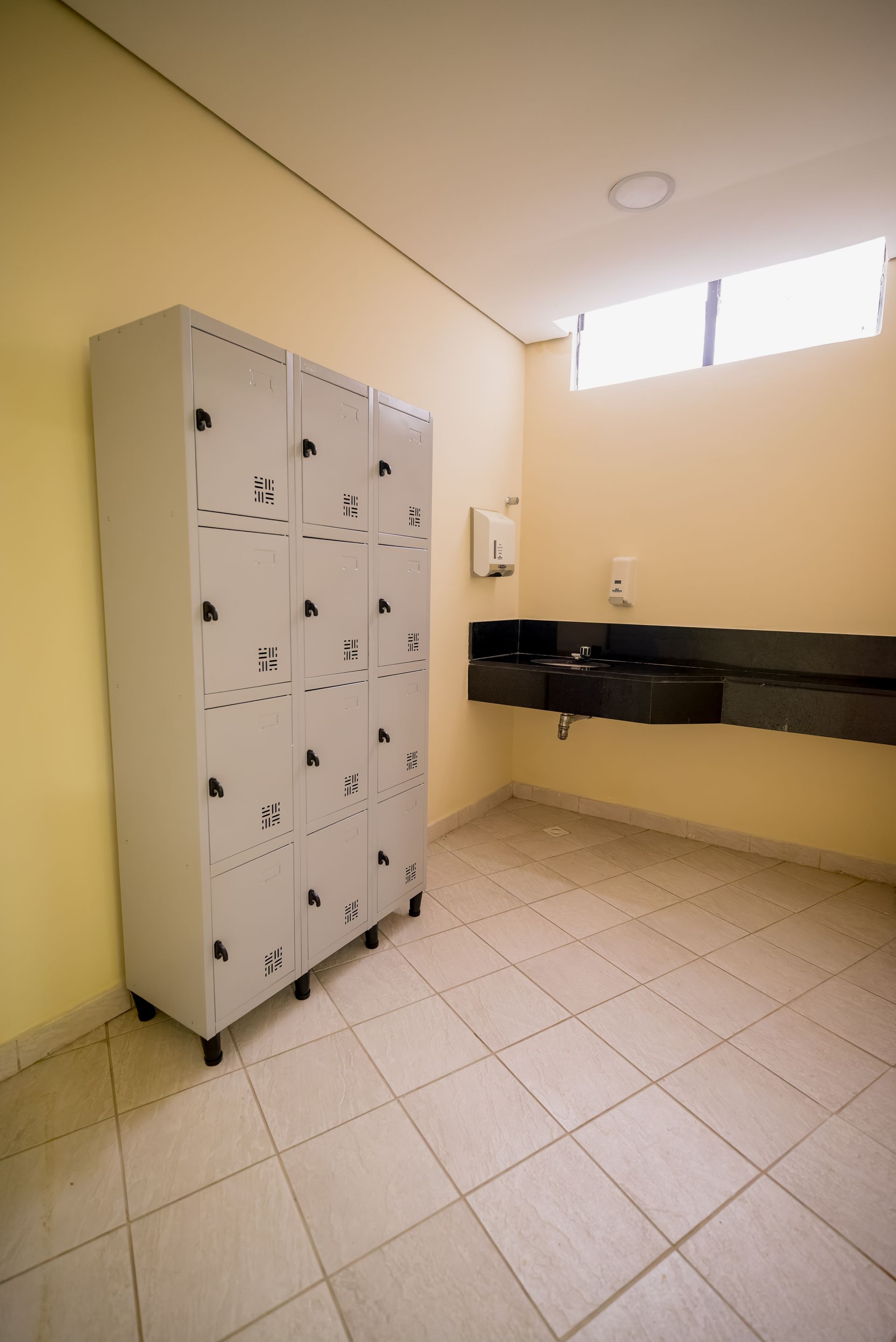 Interior de vestiário com paredes amarelo-claras, piso de azulejo e uma fileira de armários cinza ao lado de uma área com pia.