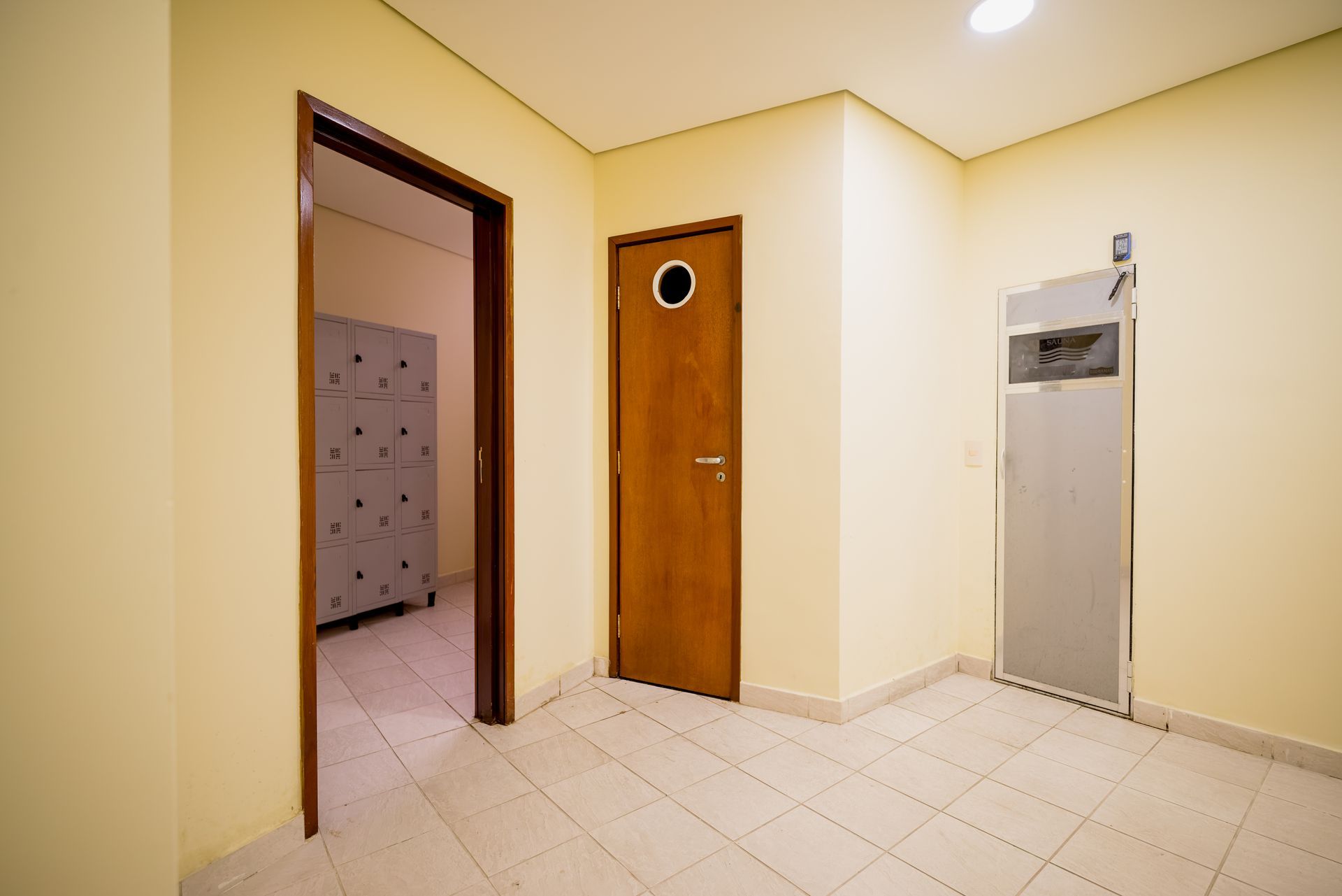 Corredor vazio com três portas. Paredes bege, piso de azulejo e painel metálico para instalações elétricas.