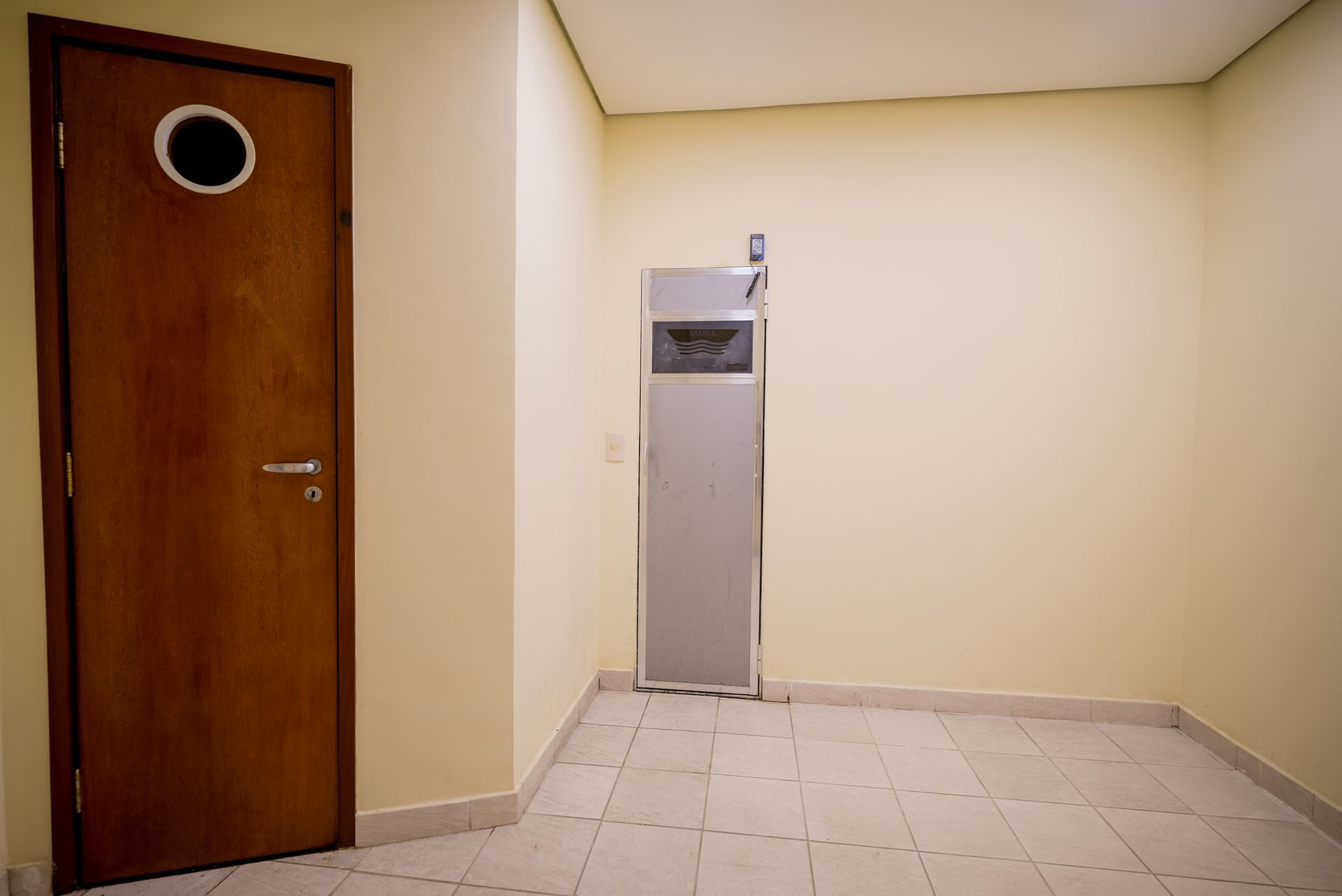 Porta marrom com vigia, eletrodoméstico de metal, paredes bege, piso de azulejo em cômodo vazio.