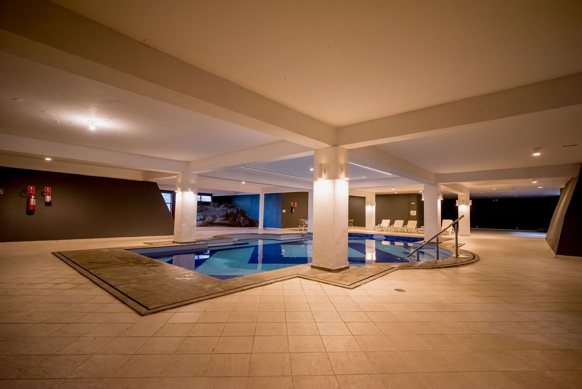 Piscina coberta com água azul e piso de azulejo bege, sob pilares e teto brancos.