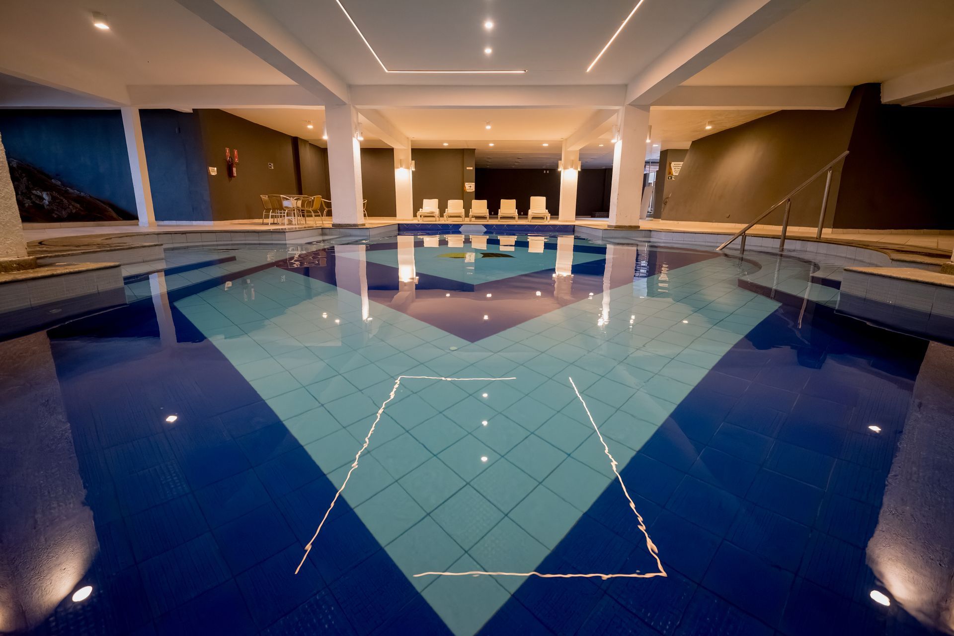 Piscina coberta com design geométrico em azulejos azuis e verde-água, luminárias espelhadas no teto e cadeiras ao fundo.