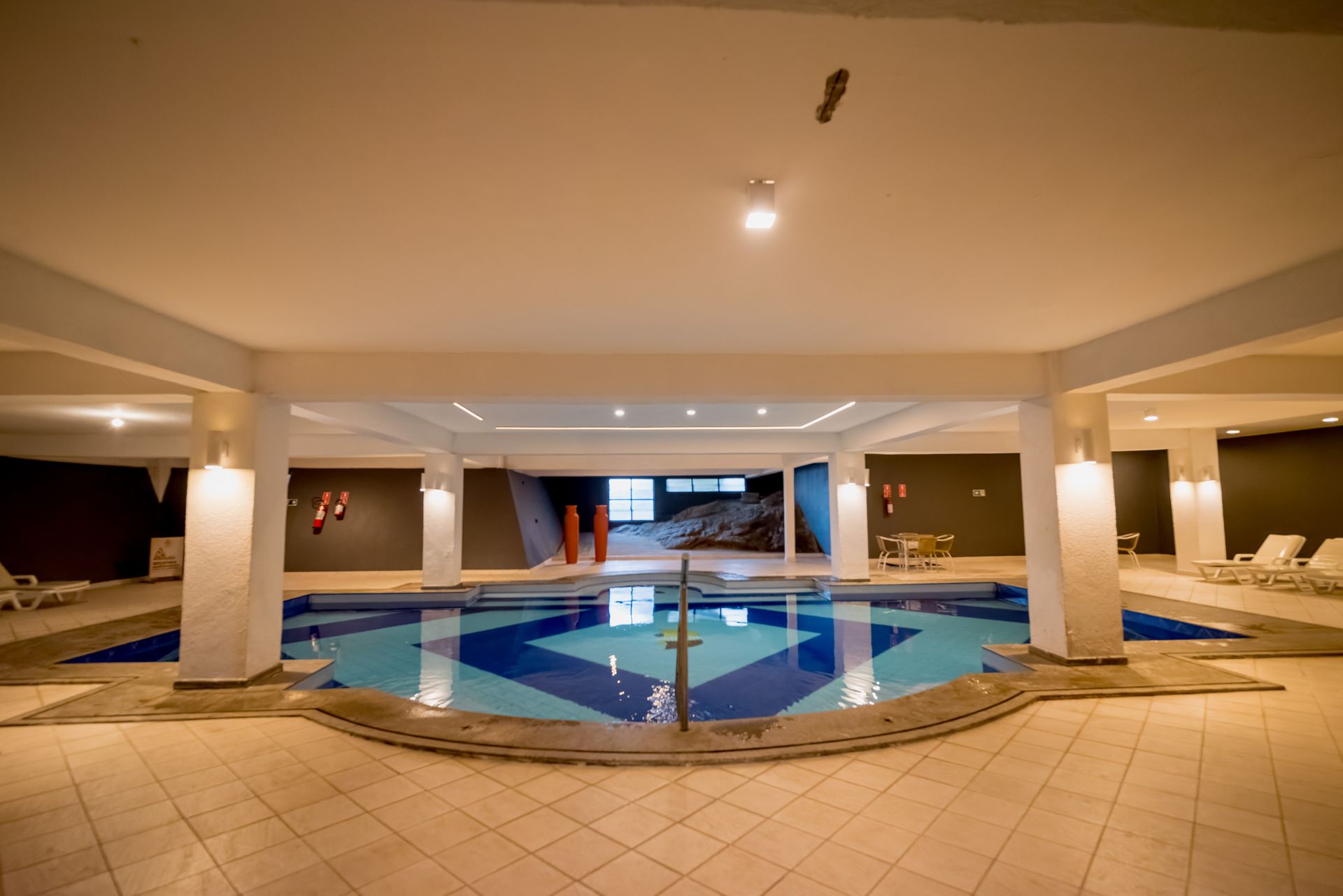 Piscina coberta com design em azulejos azuis e brancos, pilares brancos e espreguiçadeiras.