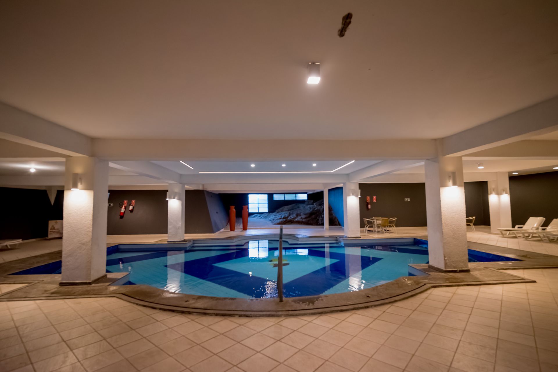 Piscina coberta com design geométrico em azulejos azuis e brancos, pilares e teto em tons neutros.