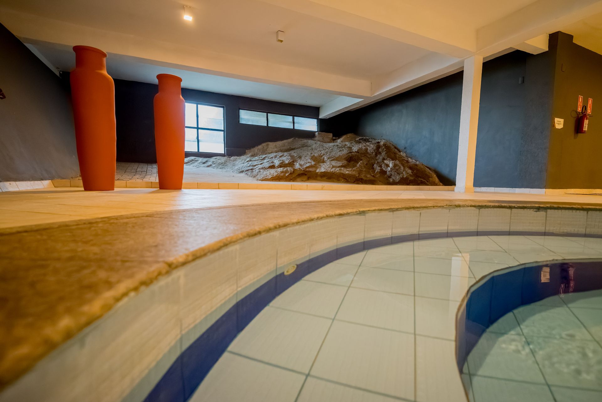 Piscina coberta com bordas curvas, piso de azulejo e dois vasos laranja; uma formação rochosa ao fundo.