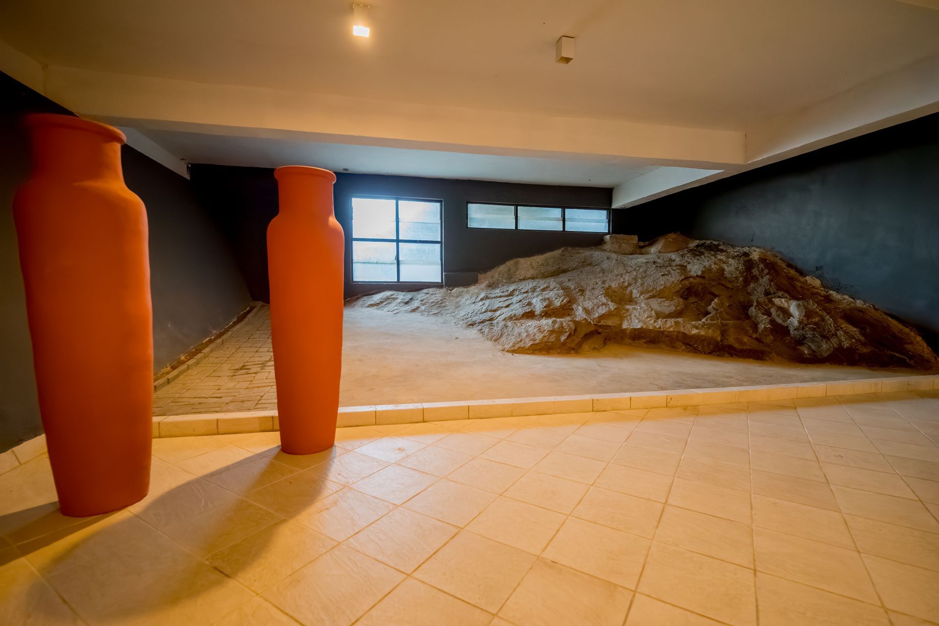 Em uma exposição de museu, dois vasos altos, de cor laranja, ladeiam uma estrutura rochosa.