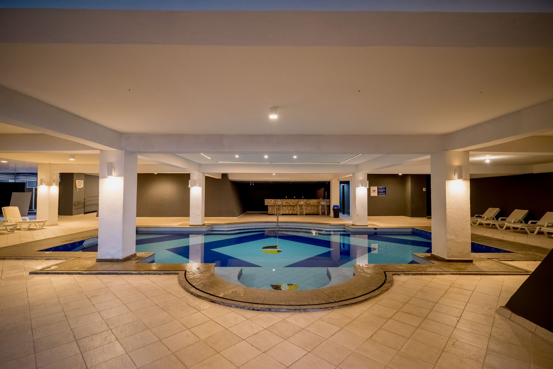 Piscina interior com azulejos azuis e estampados, rodeada por pilares brancos e piso bege.