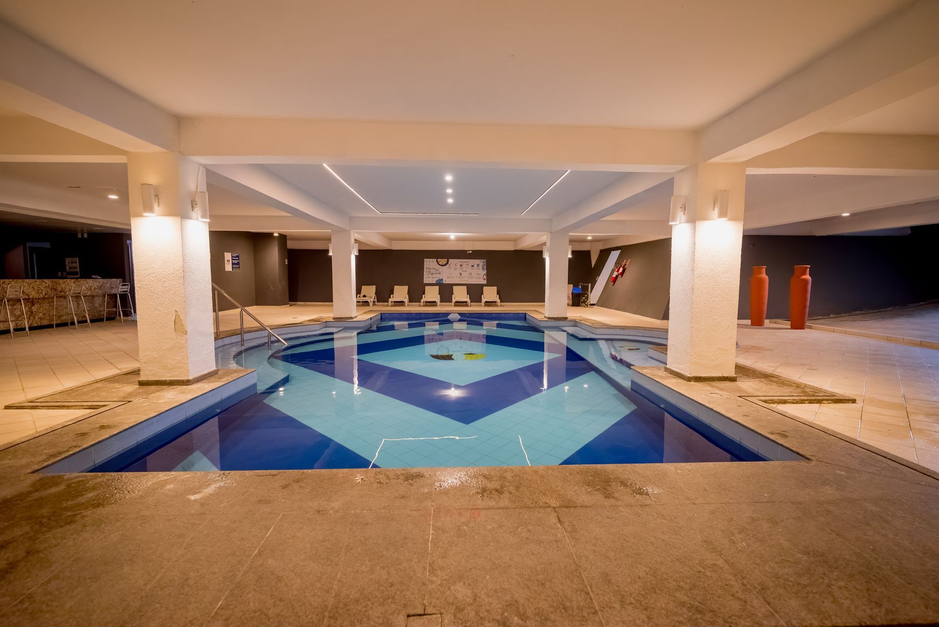 Piscina coberta com design em azulejos azuis e brancos, sob pilares brancos e iluminação no teto.