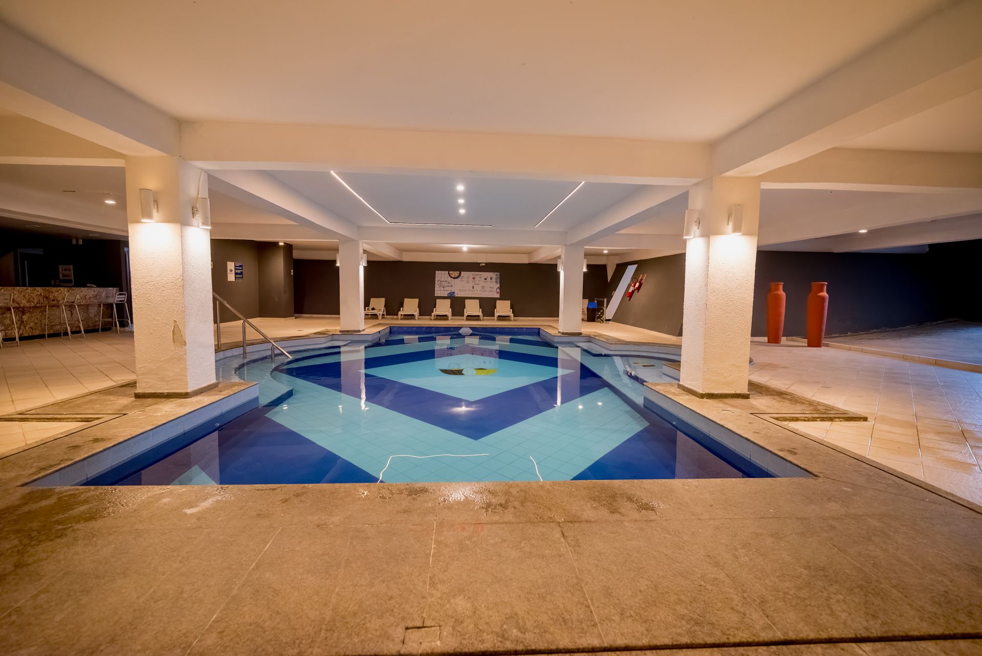 Piscina coberta com design em azulejos azuis e brancos, pilares e deck em tons neutros.