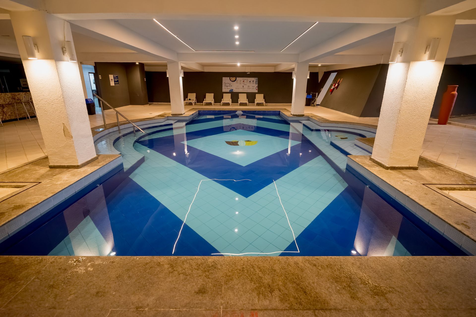 Piscina coberta com azulejos geométricos azuis, pilares brancos e espreguiçadeiras.