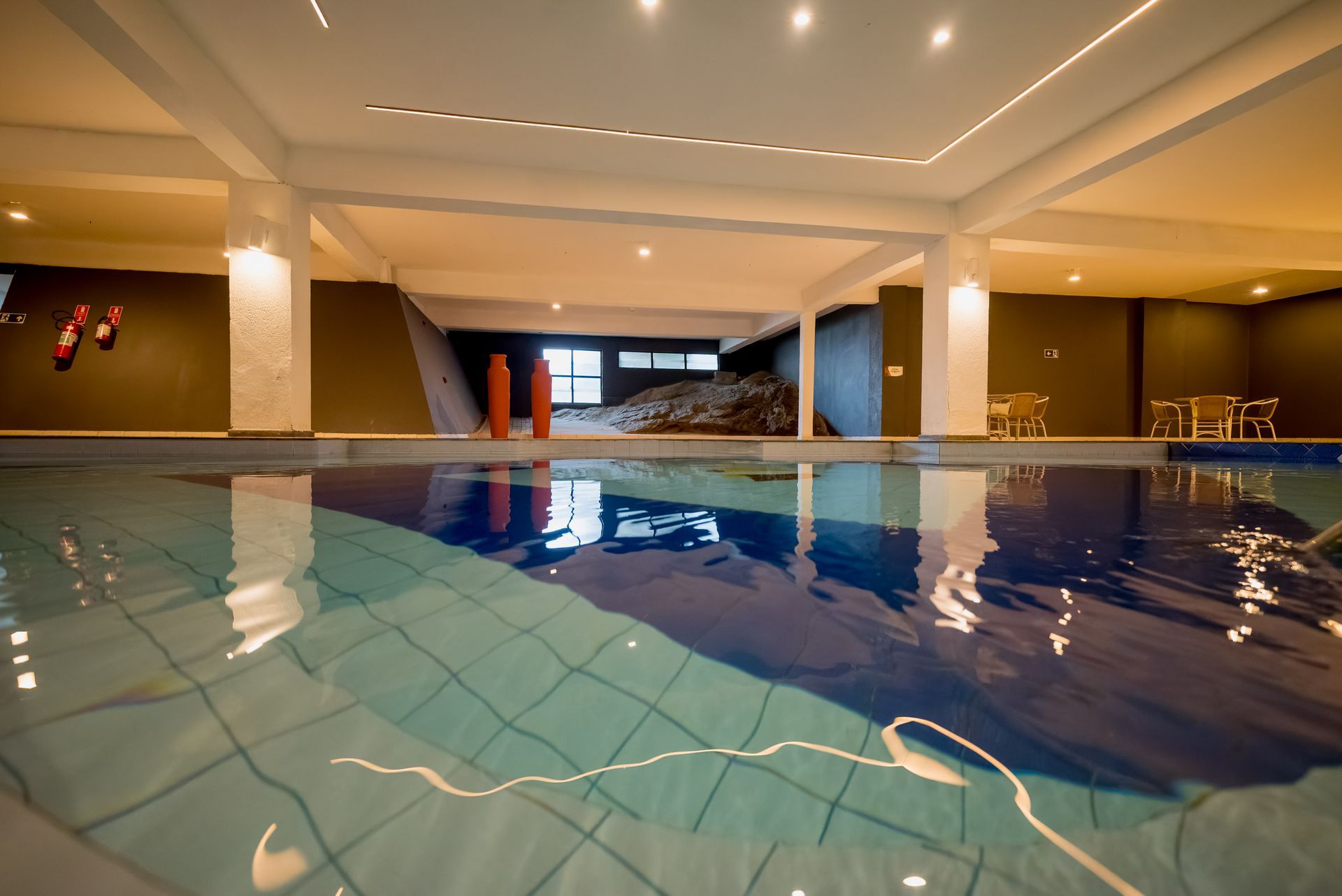 Piscina coberta com água turquesa, pilares brancos e paredes cinzentas.