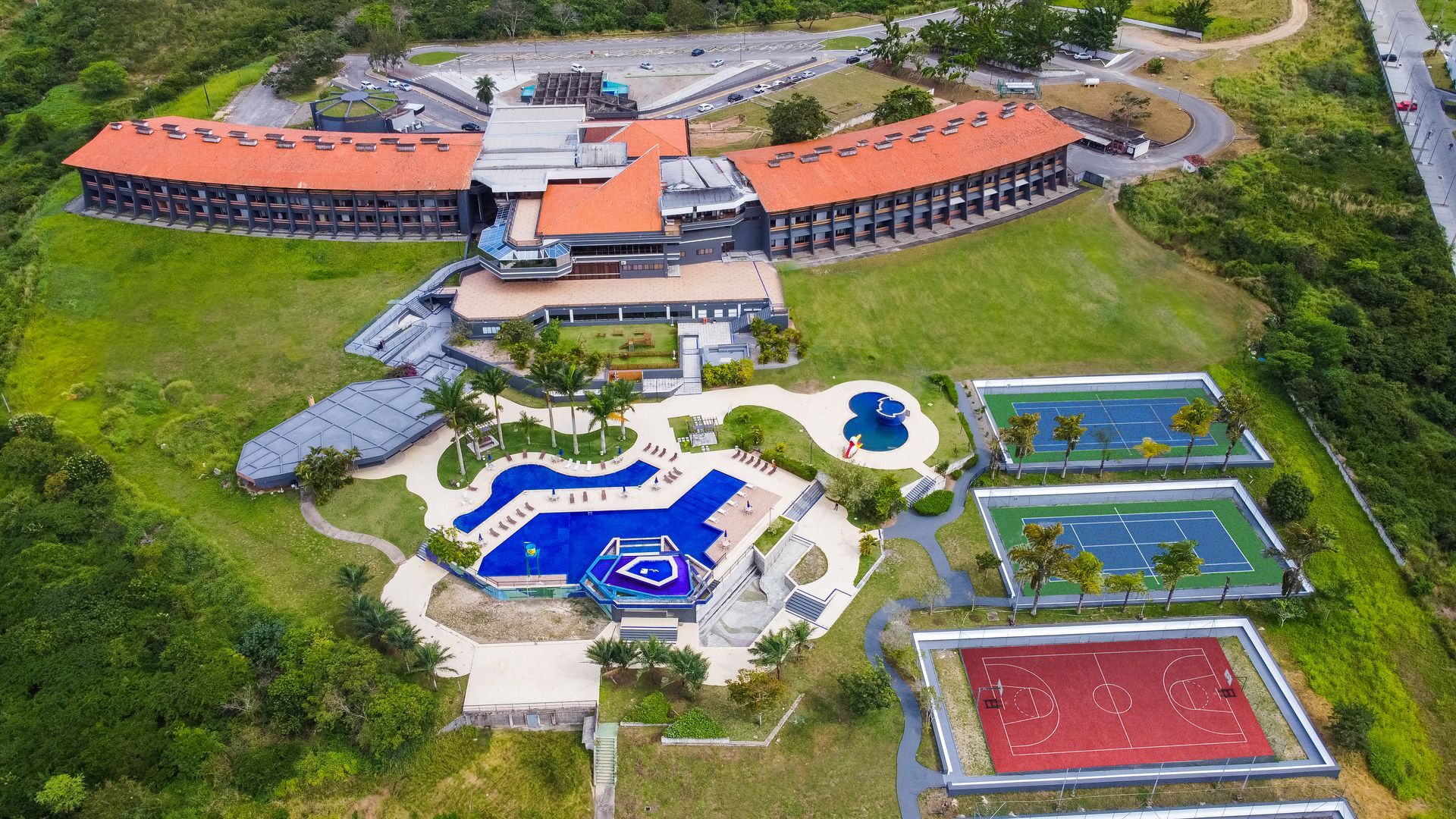 Uma vista aérea de um grande edifício com piscina e quadras de tênis cercado por árvores.
