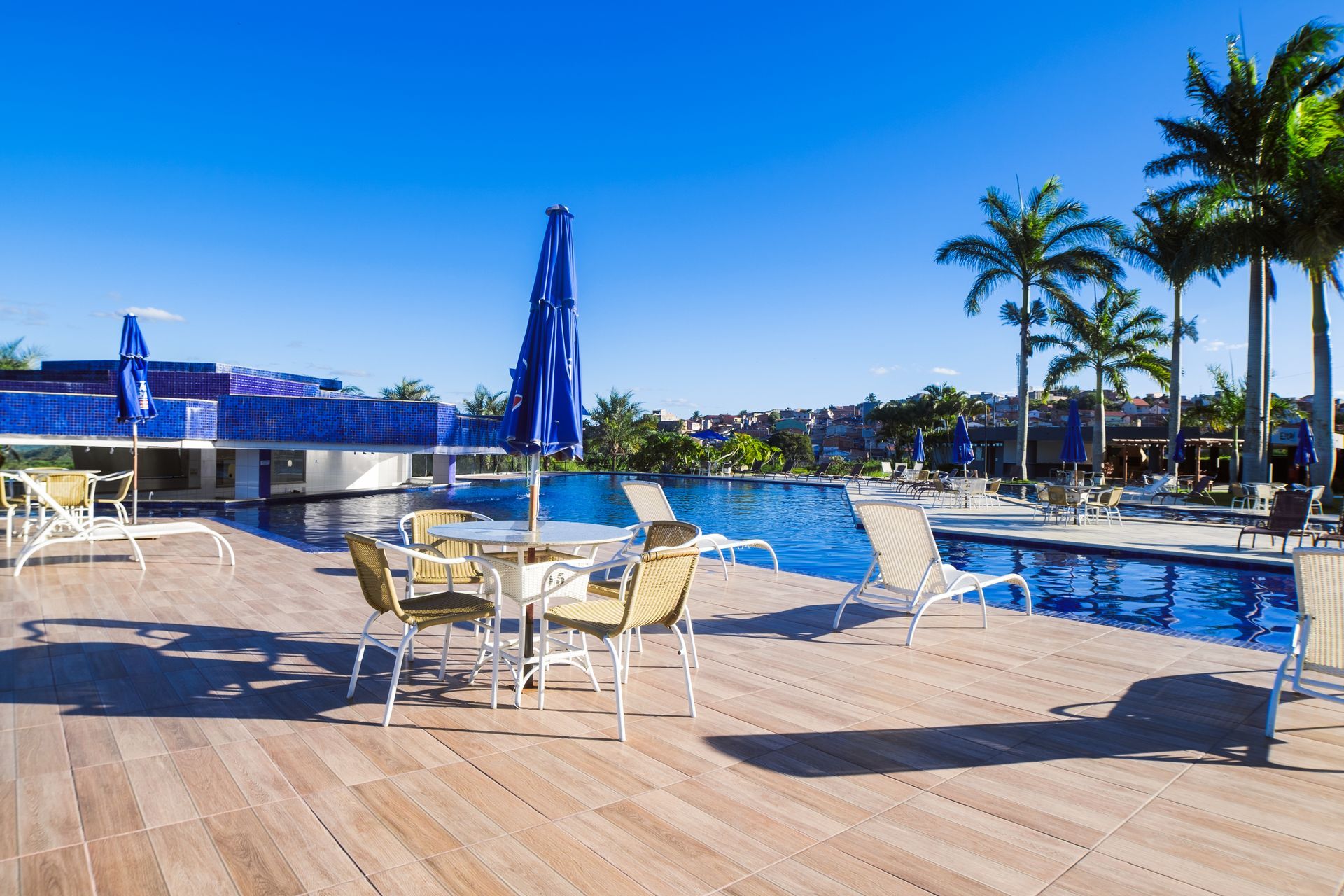 Pátio à beira da piscina com mesas e cadeiras, guarda-sóis azuis e palmeiras sob um céu ensolarado.