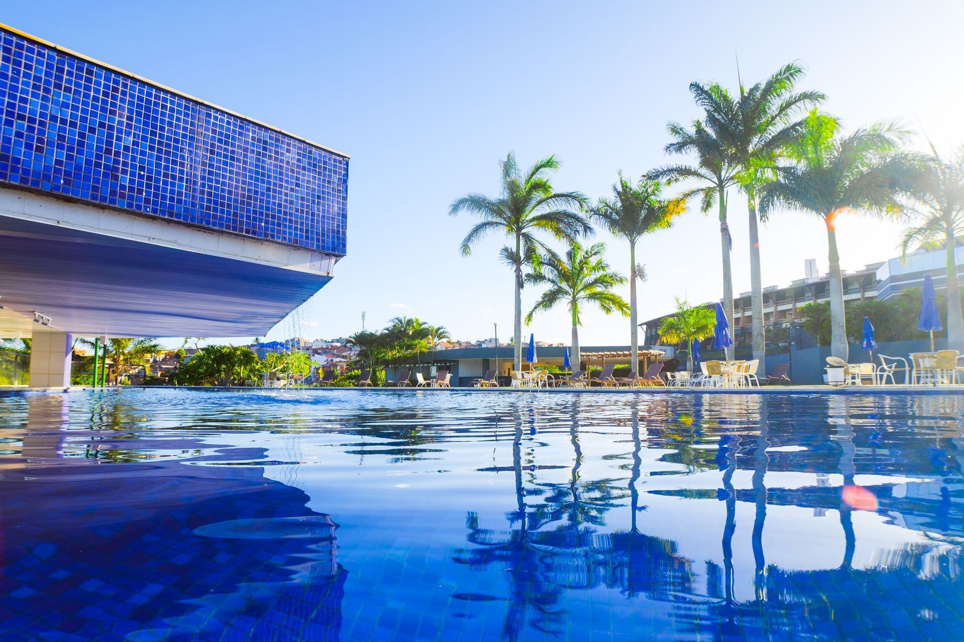 Edifício com azulejos azuis e palmeiras refletidos na piscina de um resort sob um céu claro.