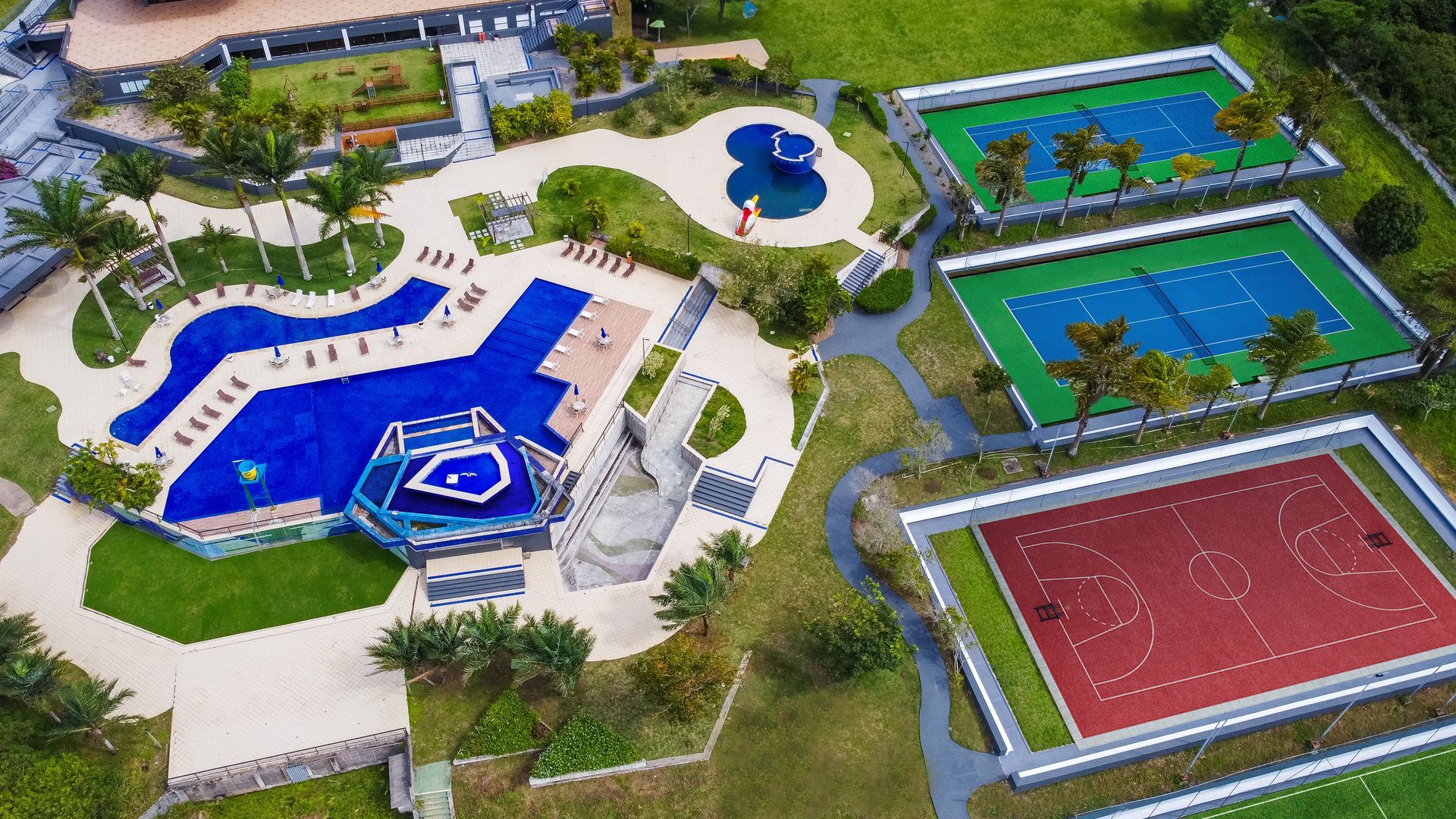 Vista aérea de um resort com piscinas azuis, quadras de tênis e uma quadra poliesportiva, cercado por vegetação.