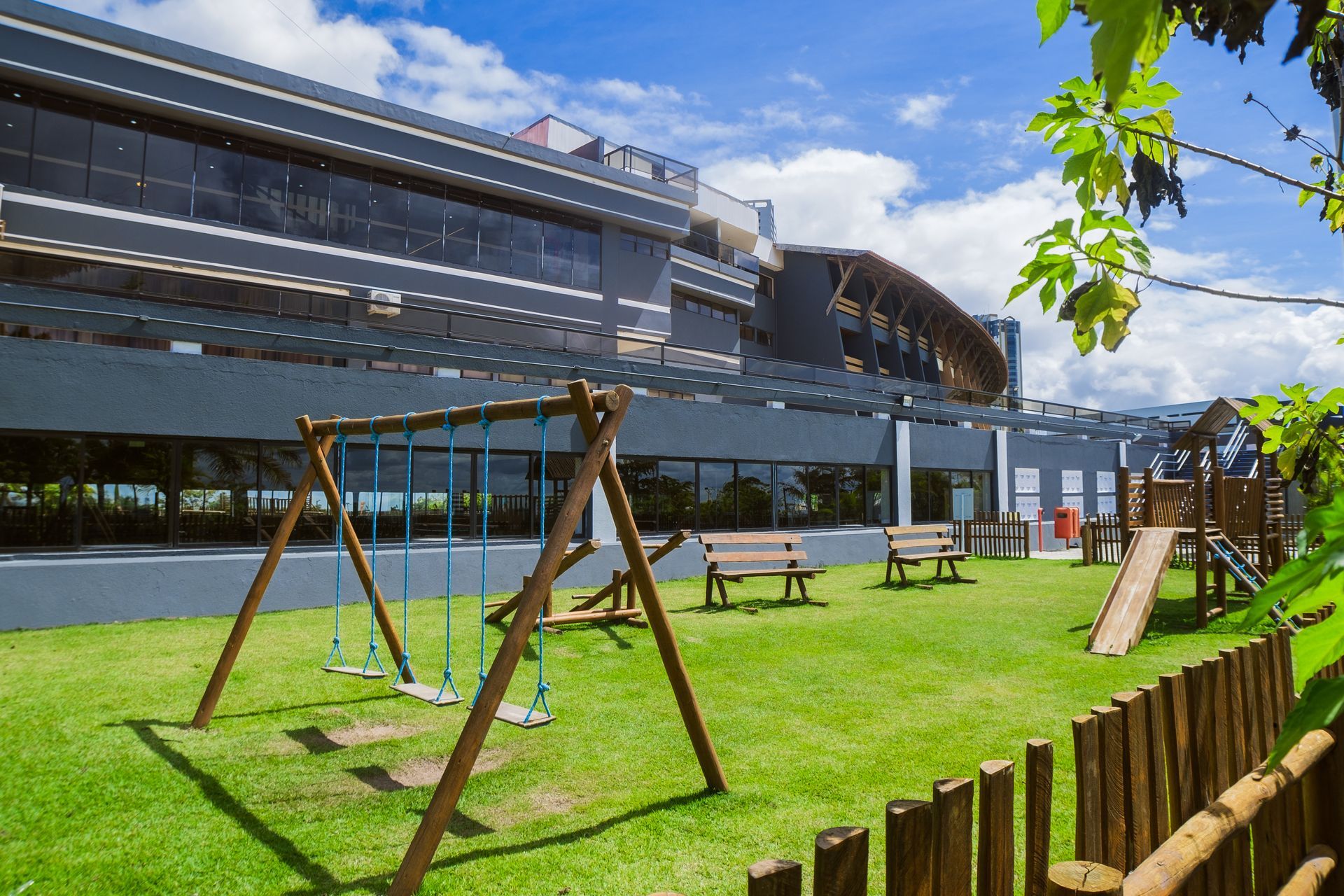 Parque infantil com balanços, escorregadores e bancos em frente a um prédio moderno com grandes janelas, em um dia ensolarado.