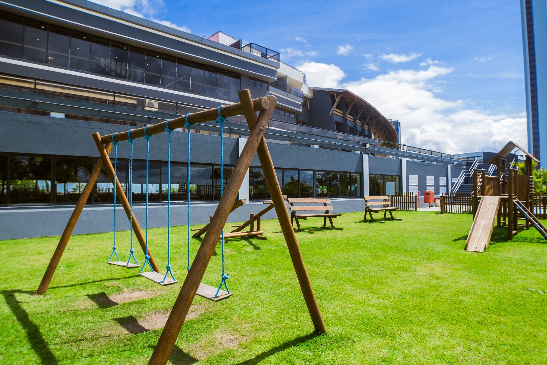 Parque infantil com balanços, escorregador e bancos em grama verde, prédio ao fundo, céu azul.