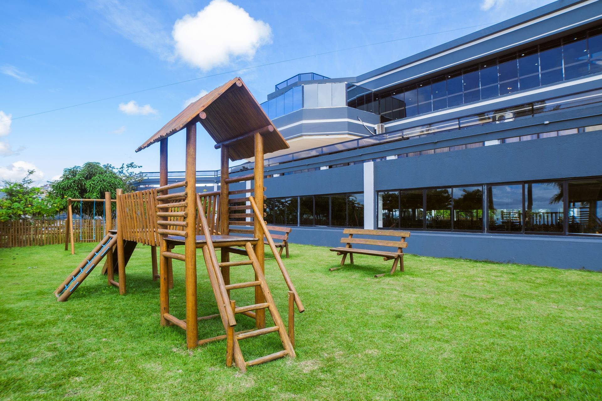 Parque infantil de madeira sobre grama verde em frente a um prédio moderno com grandes janelas.