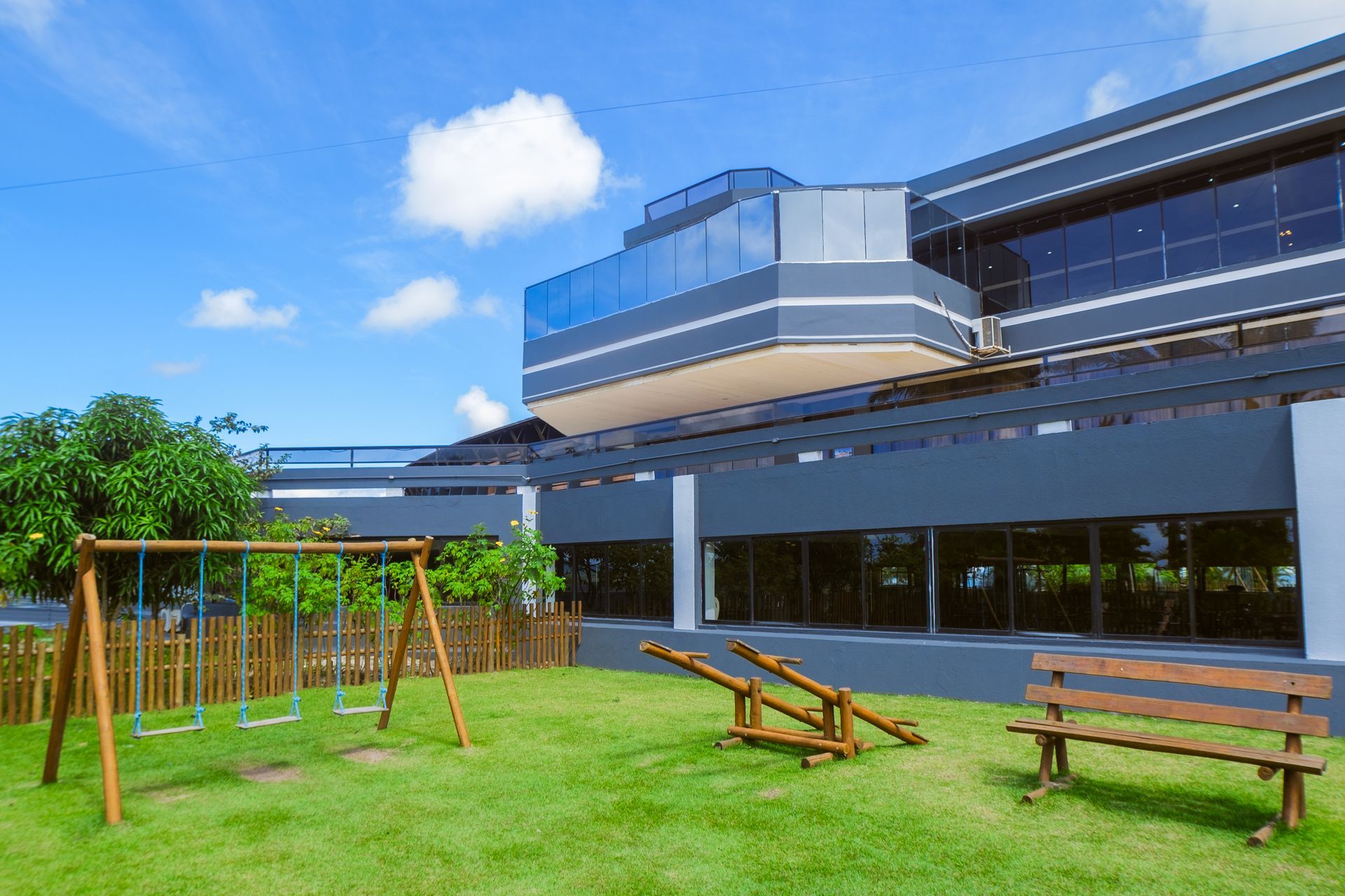 Um prédio moderno com um parque infantil equipado com balanços, gangorra e banco, sob um céu azul.