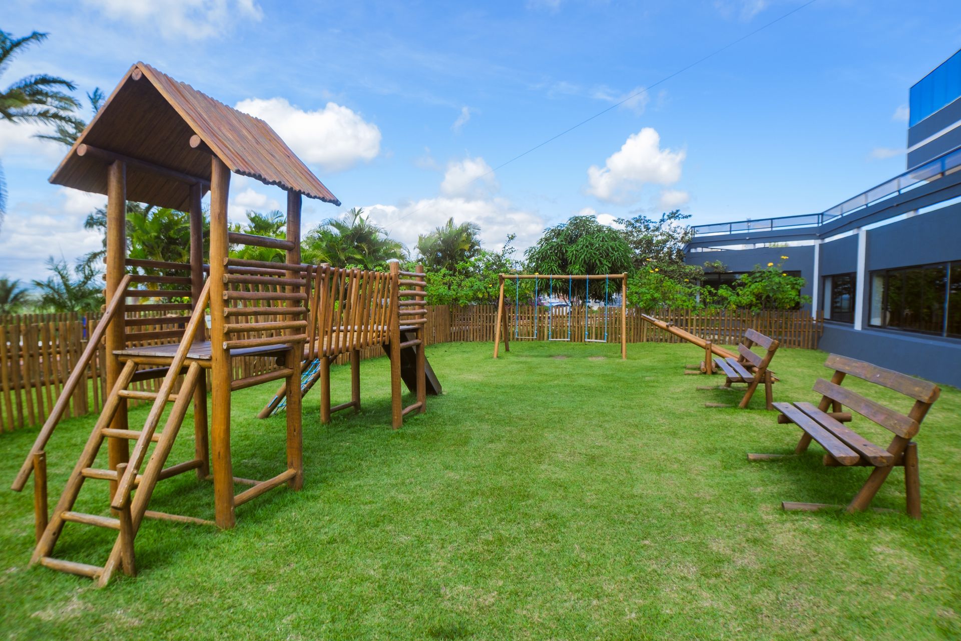 Parque infantil de madeira com escorregador, estrutura de escalada e balanços em gramado verde; bancos e prédio ao fundo.