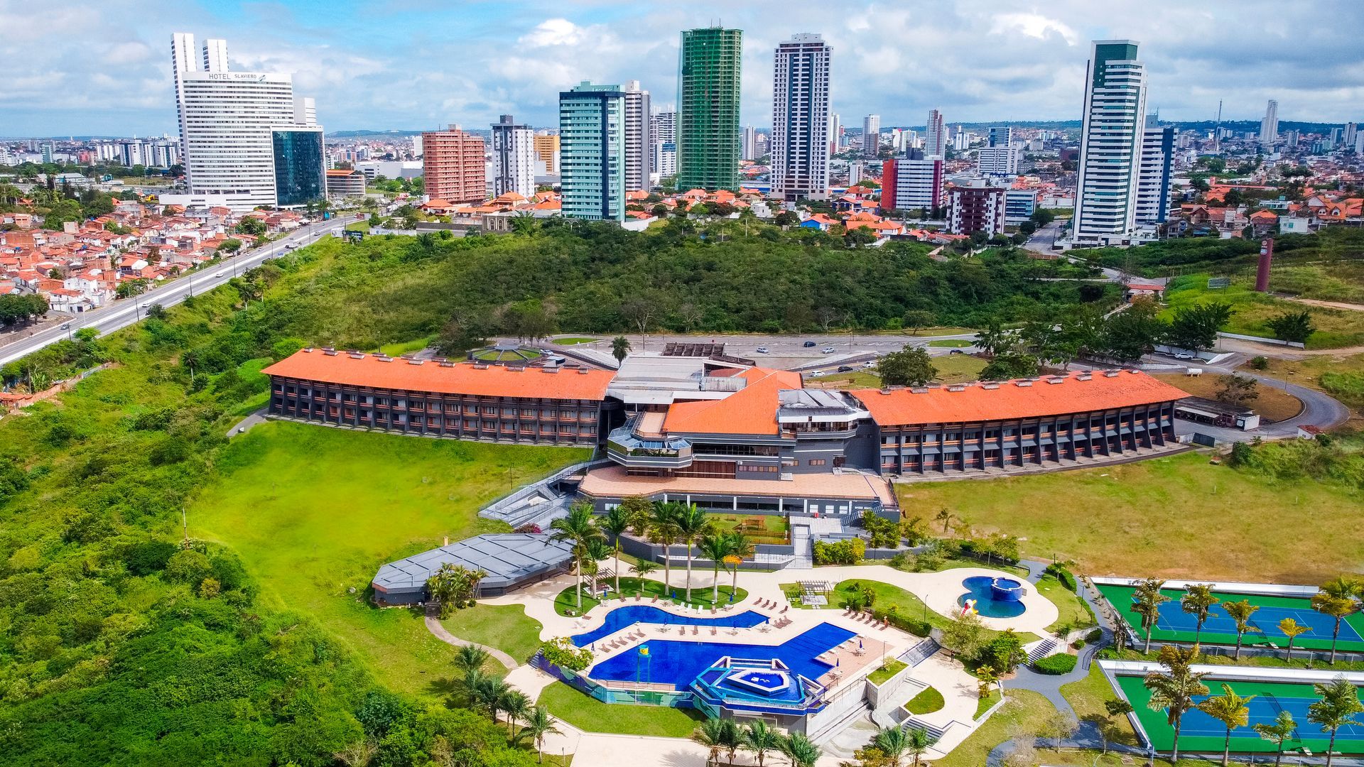 Complexo hoteleiro com piscinas e arranha-céus ao fundo, situado em uma colina verdejante.