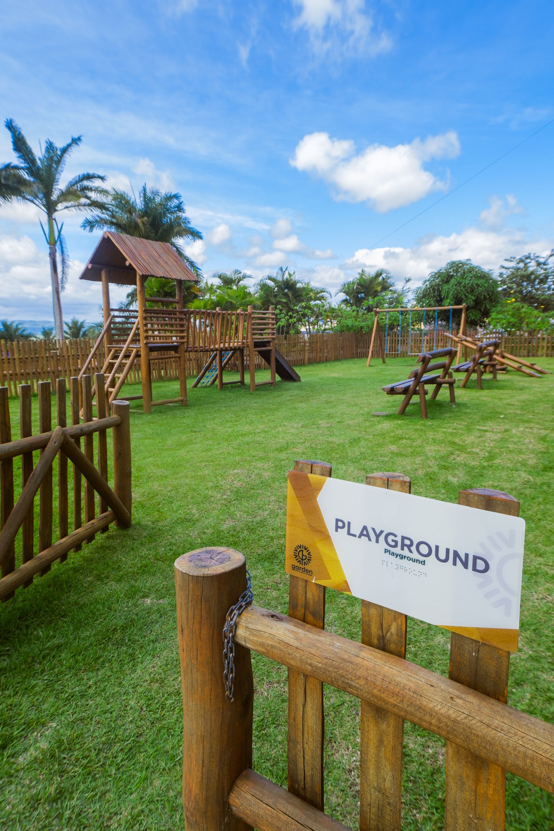 Parque infantil com estruturas de madeira, grama verde e céu azul. A placa diz 