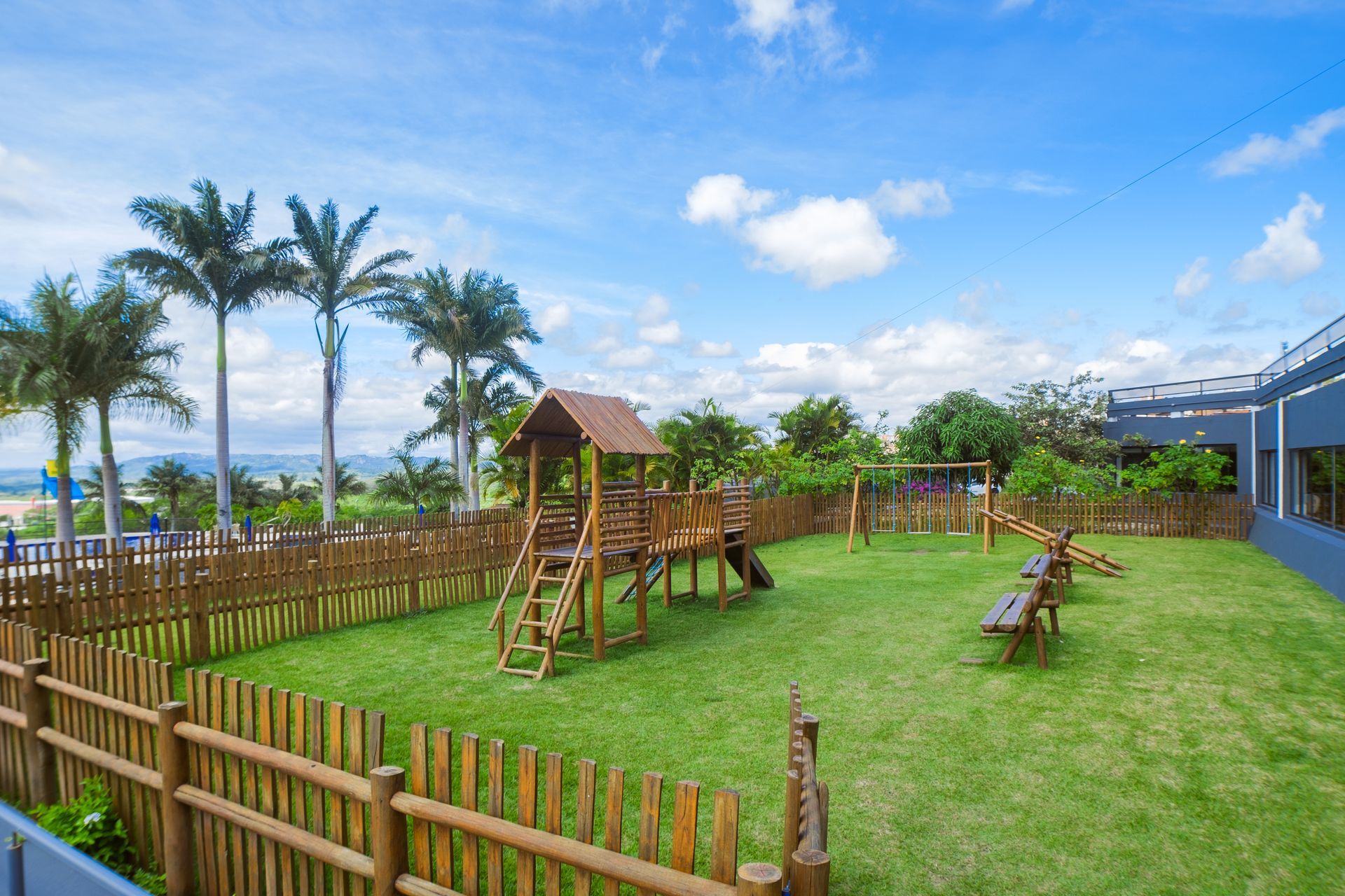 Parque infantil com estruturas de madeira, grama verde, cerca marrom e palmeiras sob céu azul.