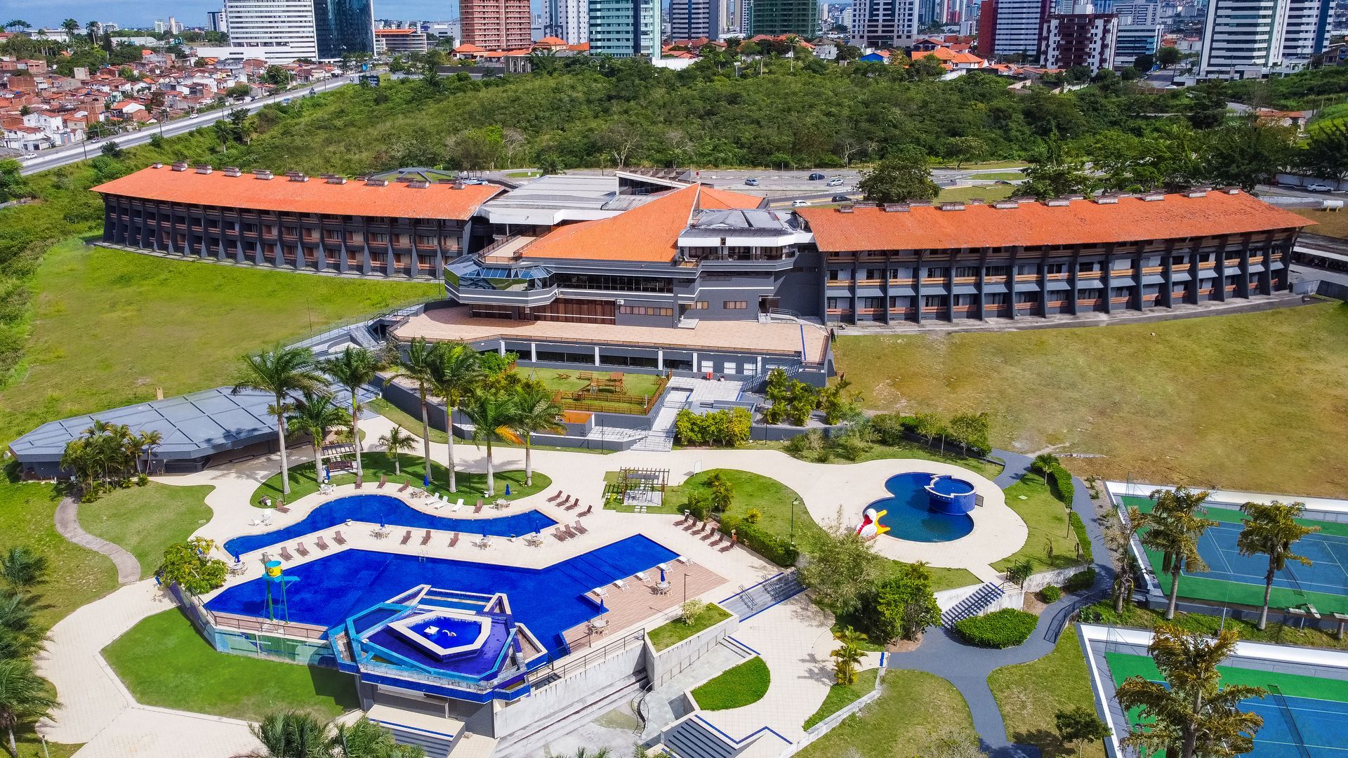 Complexo hoteleiro com piscinas, quadras de tênis e vista para a cidade. Telhado marrom, paisagem verde, água azul.