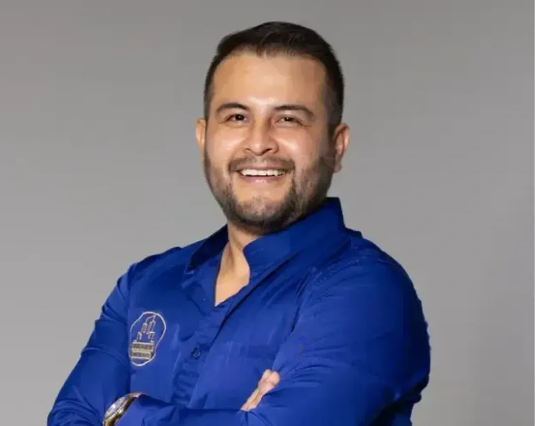 A man with a beard is wearing a blue shirt and smiling with his arms crossed.