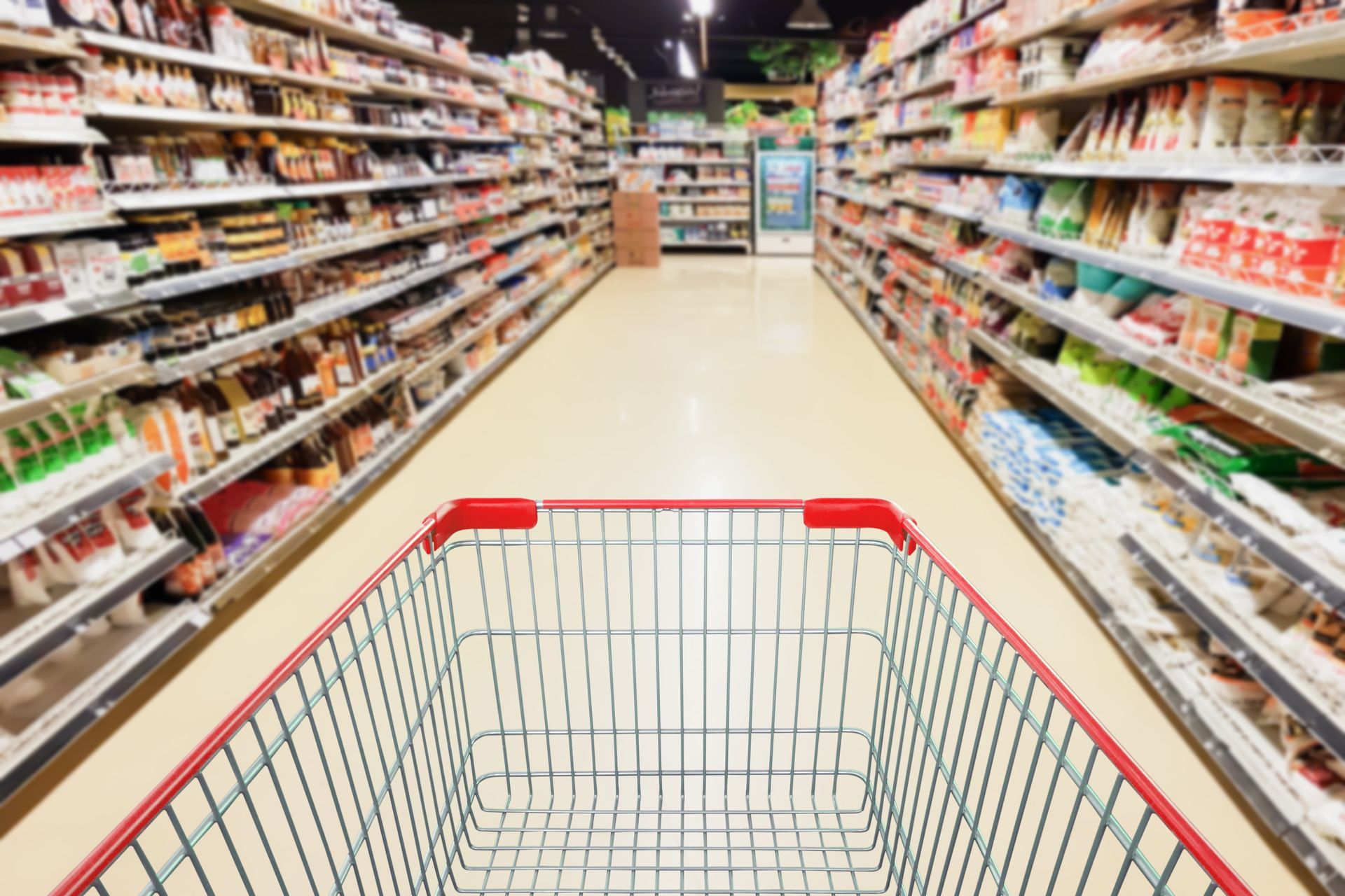 Empty red shopping cart in grocery store aisle filled with products.