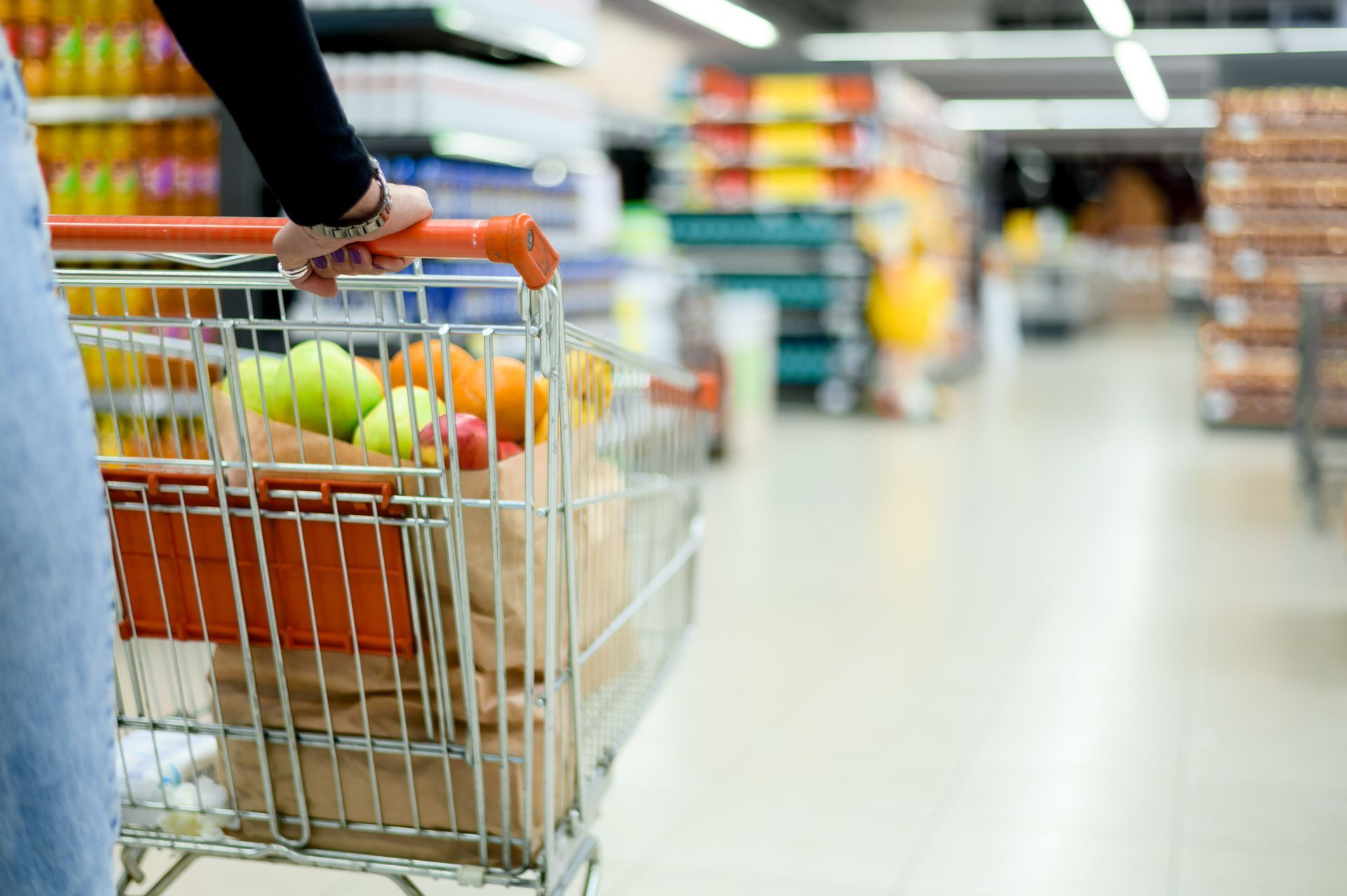 Person pushing shopping cart filled with fruit through supermarket aisle.