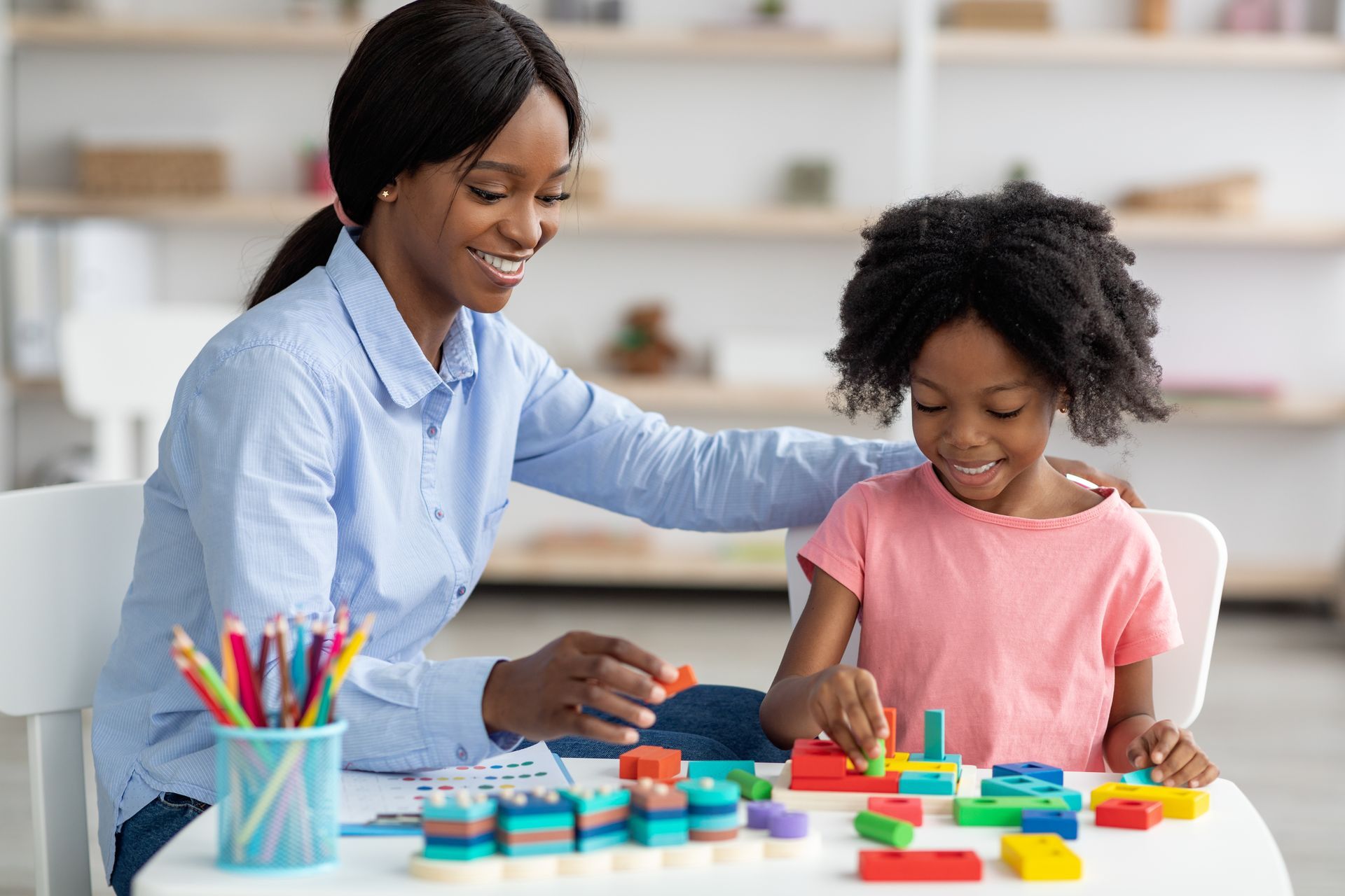 Woman assists child playing with colorful blocks at a table.