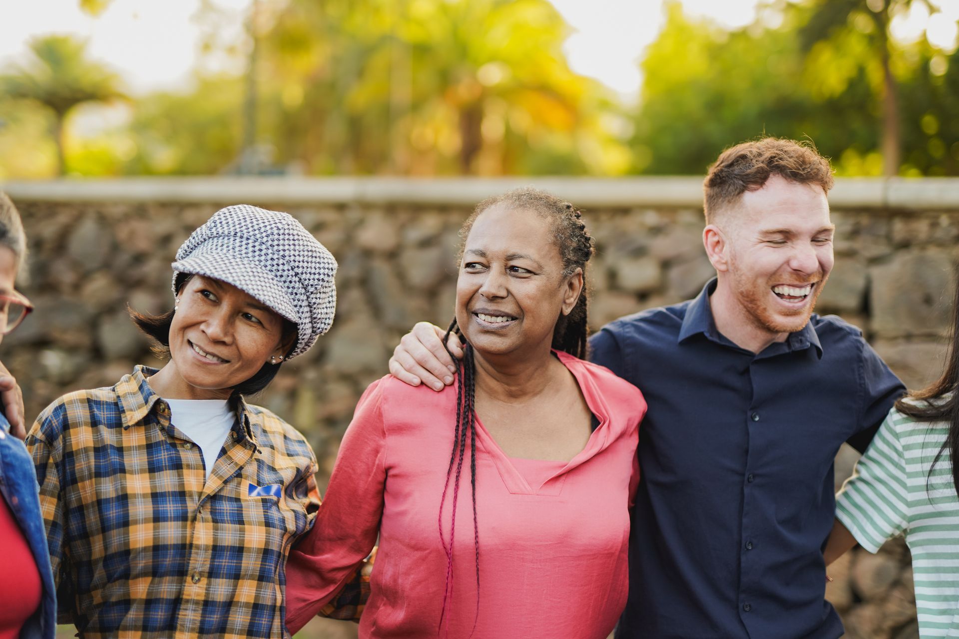 Group of people smiling, arm-in-arm outside. Brick wall and trees in background.
