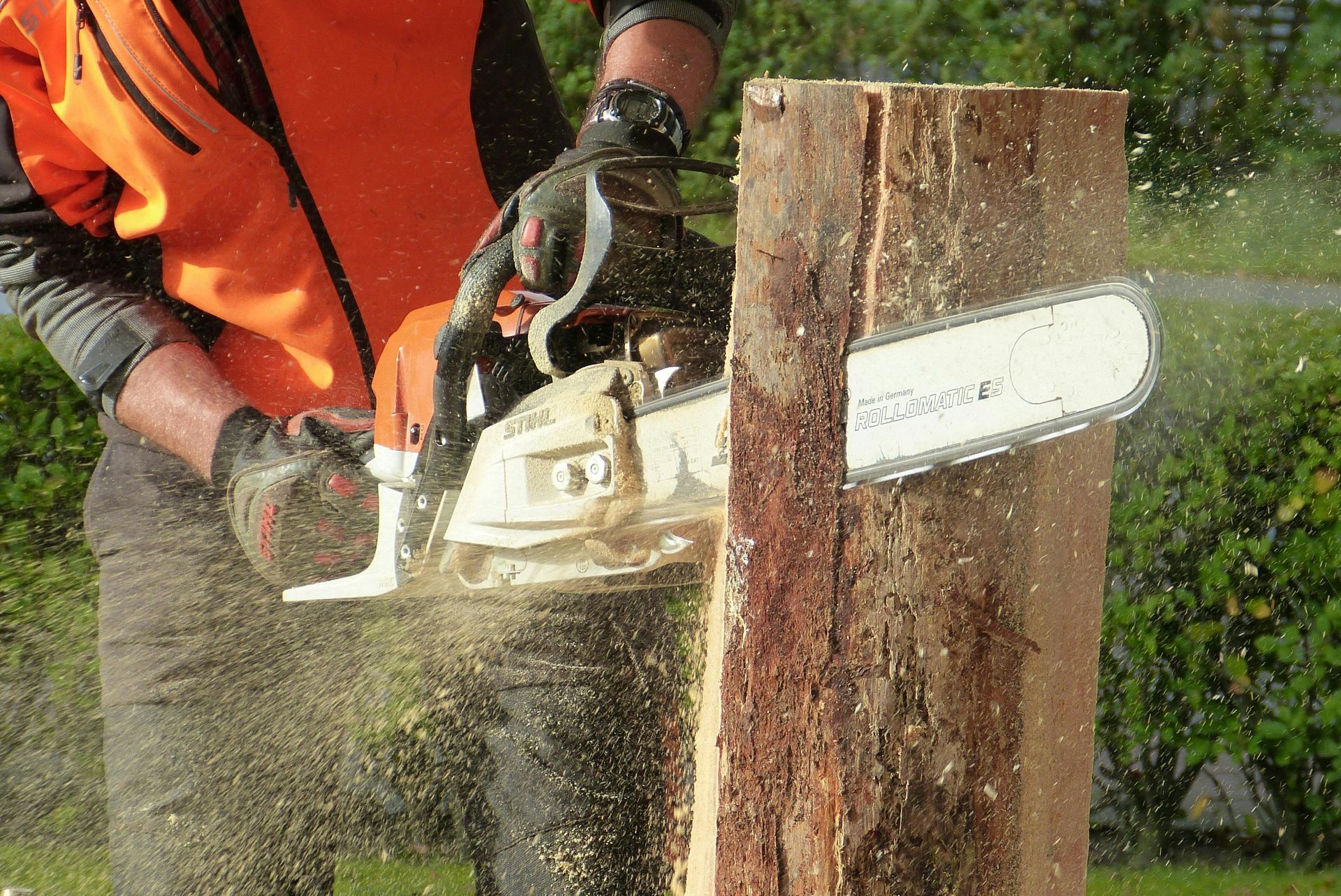 Worker in orange gear using a chainsaw to cut a wooden post, with sawdust spraying.