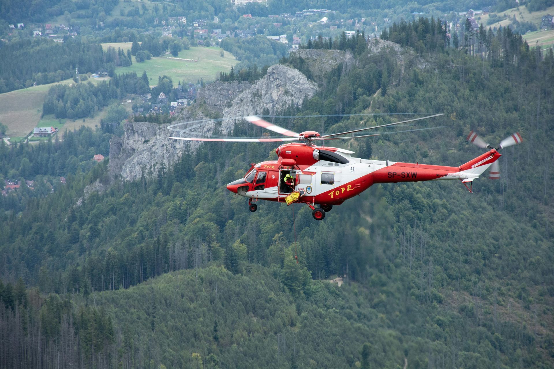 Red and white helicopter flying over green forested mountains and a valley
