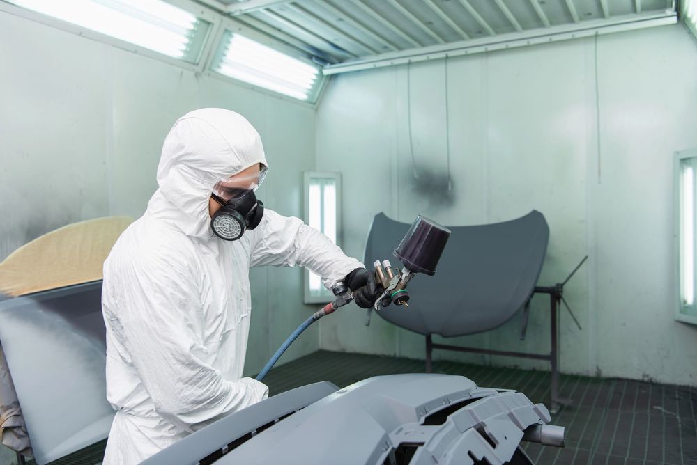 Una persona con indosso una tuta protettiva bianca vernicia a spruzzo dei componenti di un'auto all'interno di una cabina di verniciatura.