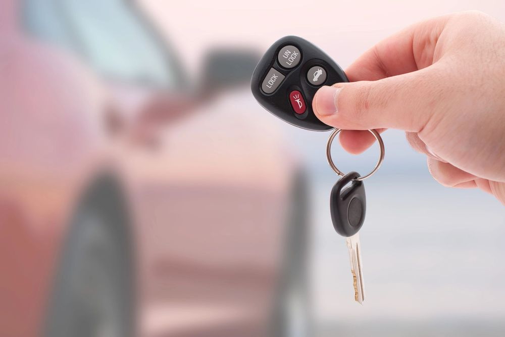 Una mano che tiene le chiavi dell'auto e un telecomando davanti a un'auto parcheggiata sfocata