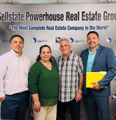 Group of four people posing in front of a real estate company logo, smiling.