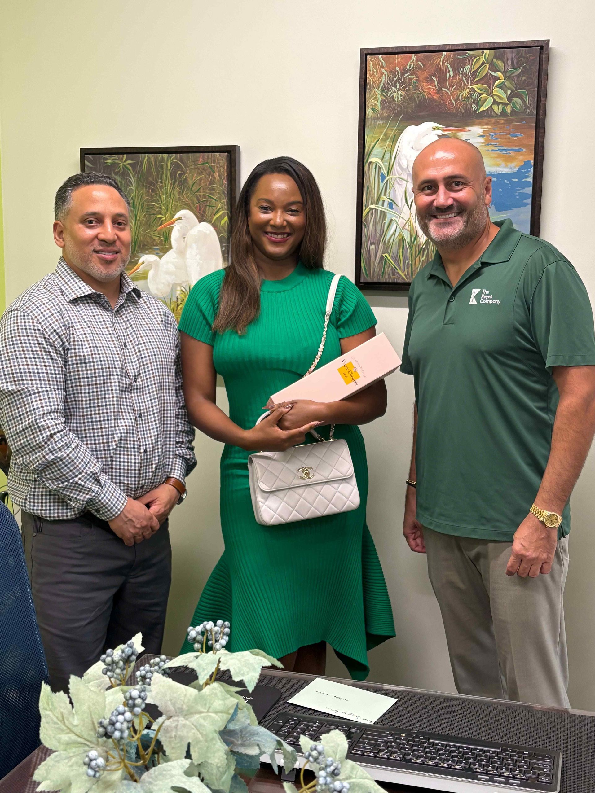 Three people pose indoors: a woman in green dress holds a pink box, flanked by two men.