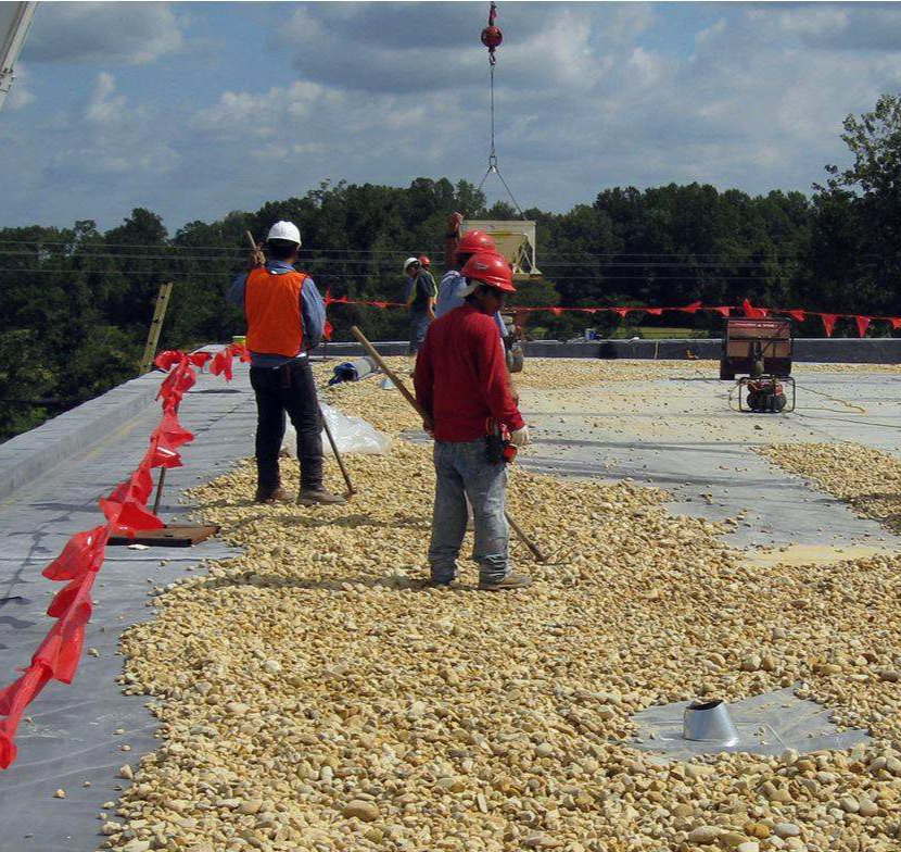 Construction workers on a flat roof, spreading gravel; crane above. Bright orange safety cones. Sunny day.