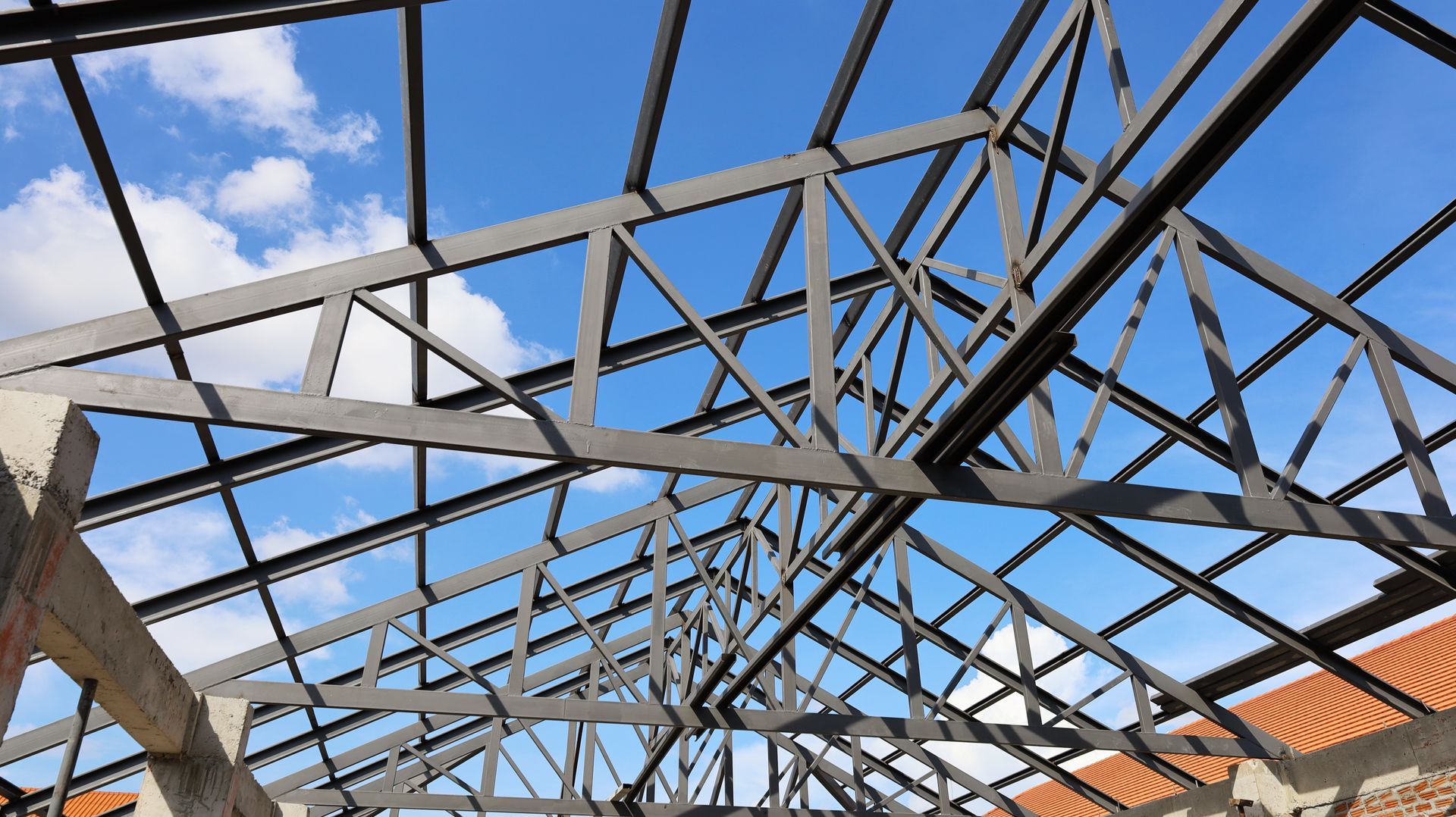 Steel roof truss framework against a blue sky with scattered clouds.
