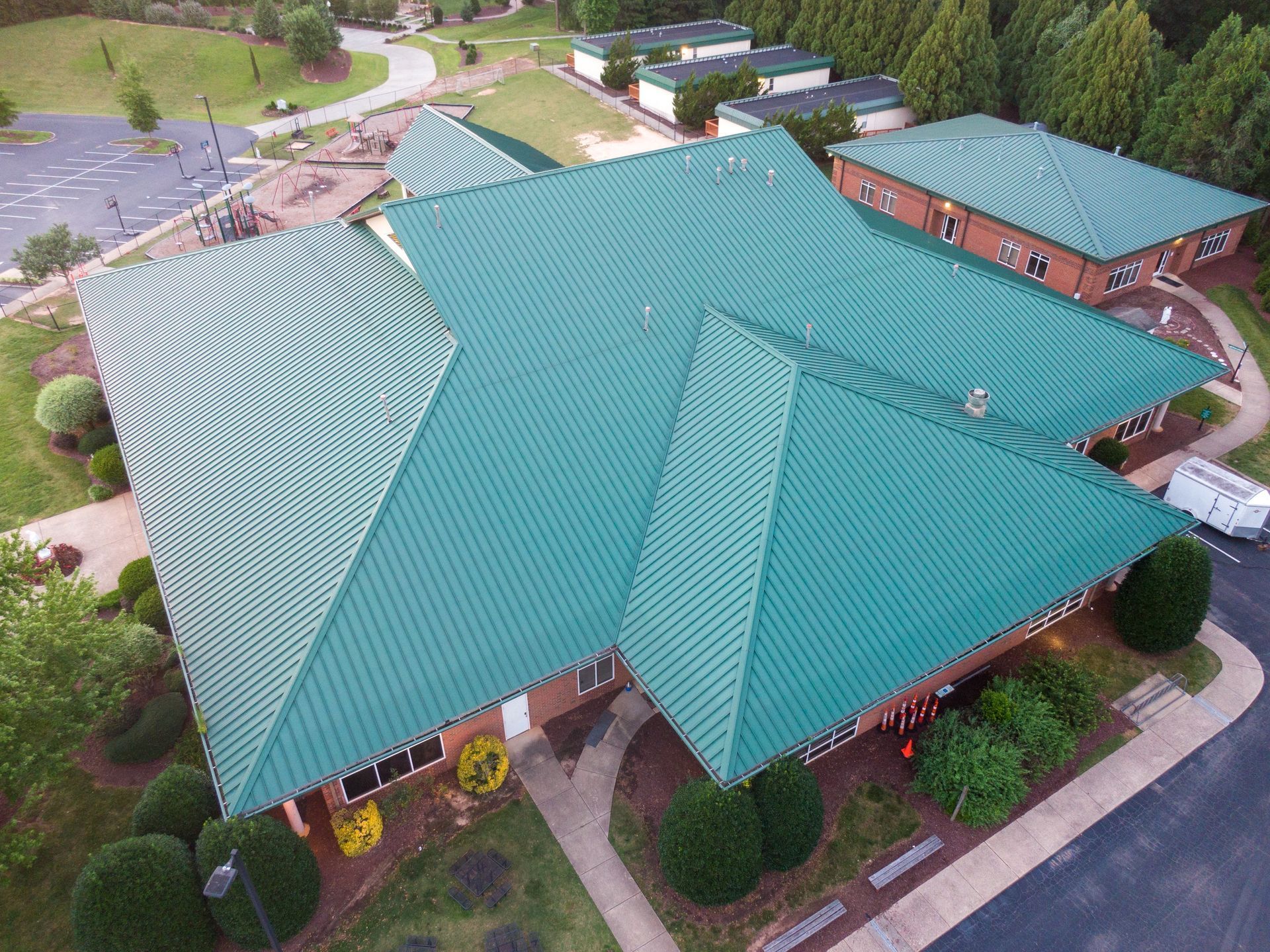 Aerial view of a building with a large green metal roof and surrounding landscaping, near a playground.